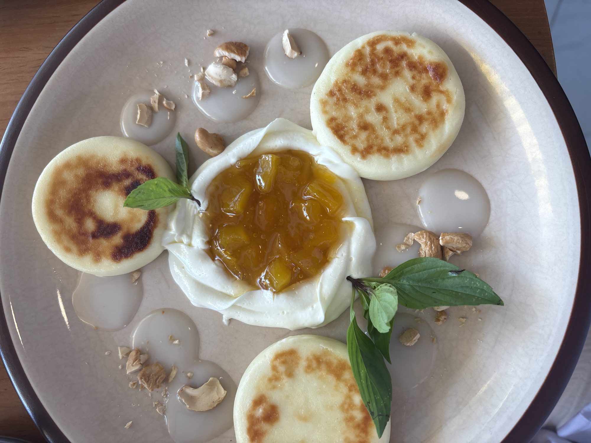 A top-down shot shows a plate containing three small, round, browned pancakes or syrniki, a dollop of white cream cheese topped with orange fruit preserves in the center, basil leaves as garnish, drops of a light sauce, and chopped nuts on a light-colored plate. The plate has a dark brown rim. The scene suggests the meal is being served in Nha Trang, Vietnam, given the presence of a Western dish prepared with possible Vietnamese ingredients.