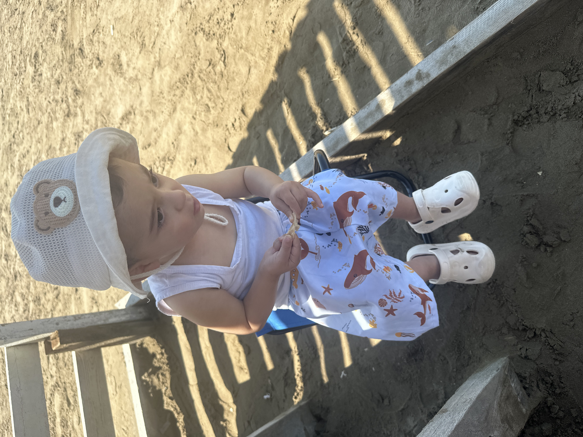 A young child, wearing a white bucket hat with a bear emblem, a white tank top, and white pants with an ocean-themed print featuring whales and starfish, sits on a blue plastic chair. The child is holding a piece of food in their hands. The child is wearing white perforated clogs. The setting appears to be a sandy area, possibly a beach or playground, with a wooden structure casting striped shadows on the sand. This scene is located in the Sabunchu Raion of Azerbaijan.
