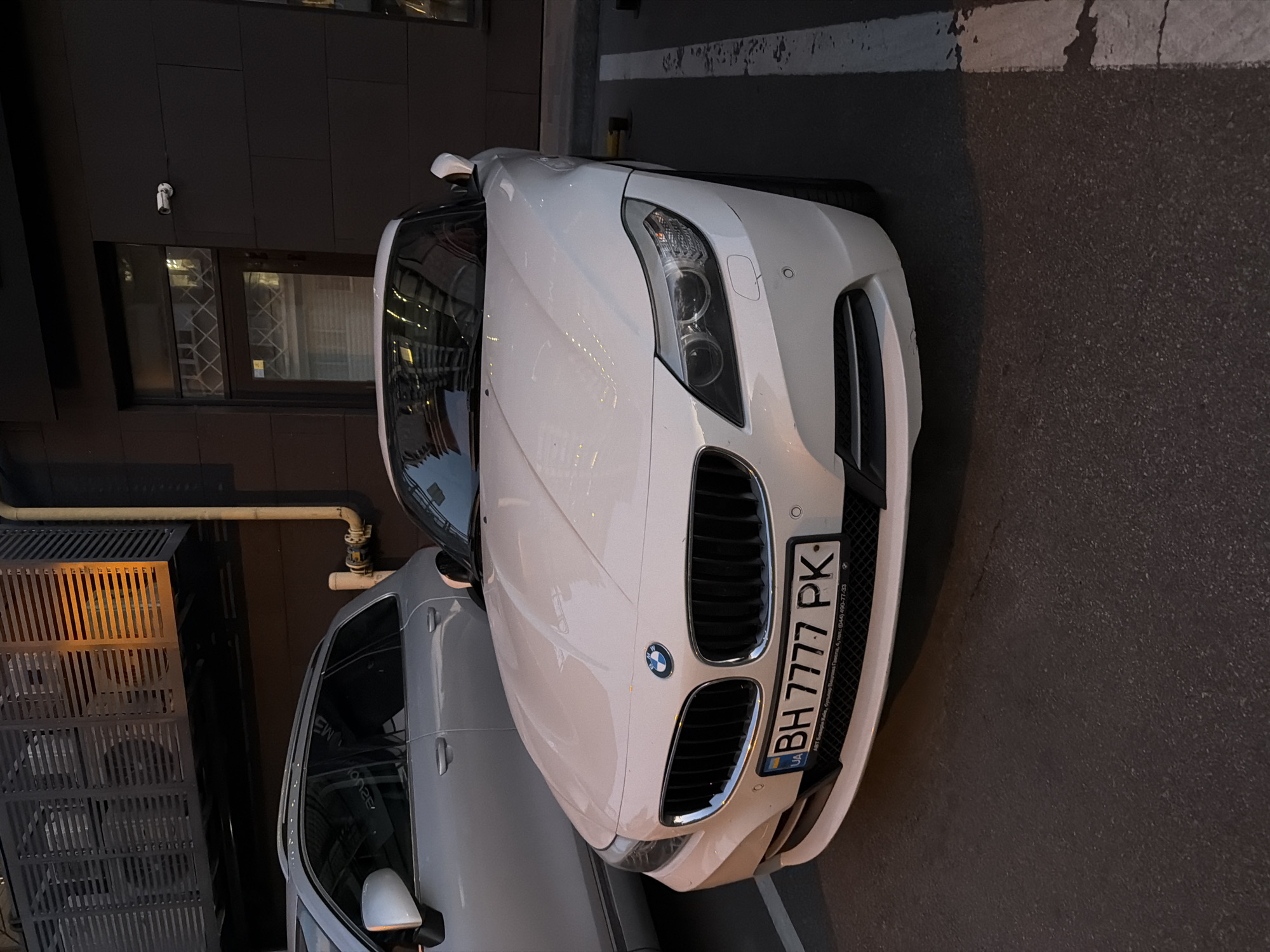 A white BMW Z4 convertible is parked in a lot, facing the camera. The car is clean and in good condition. The license plate reads "BH 7777 PK". Behind the BMW, a silver SUV is partially visible. The background consists of a dark building with a security camera mounted on the wall, and a metallic structure with orange lighting. The pavement is asphalt with white parking lines. The scene appears to be in an urban environment.