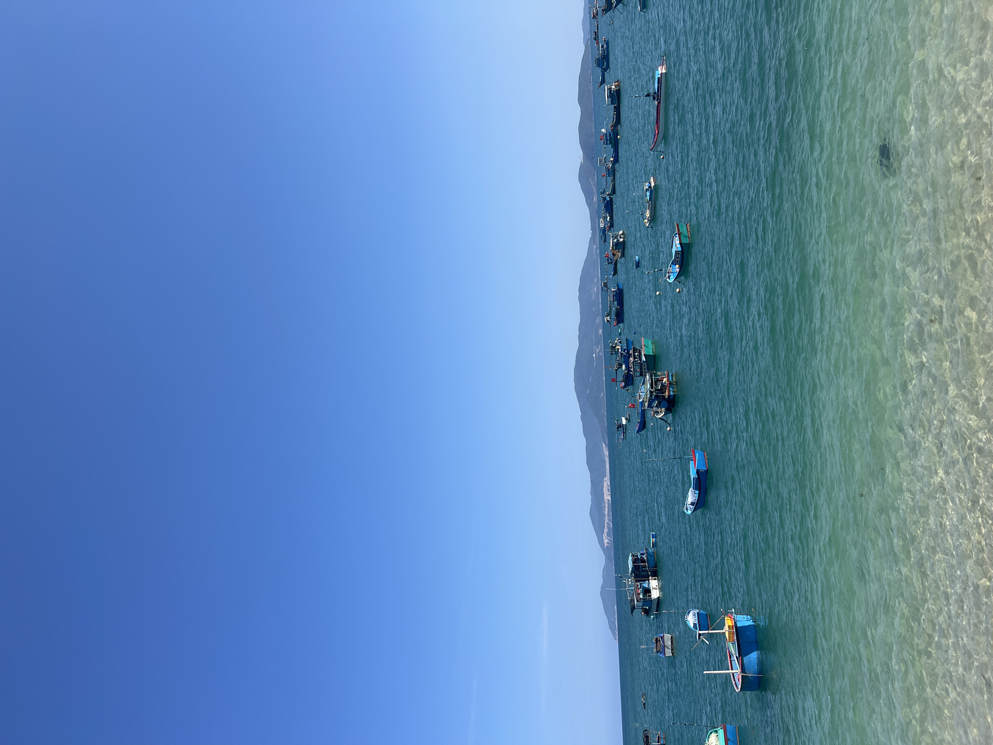The image displays a wide-angle view of a bay in Nha Trang, Vietnam, under a clear, deep blue sky that occupies the upper half of the frame. The lower half features calm, turquoise-to-emerald green ocean water. In the immediate foreground, the water is shallow and clear, revealing a rippled, light-colored sandy seabed. Dozens of traditional Vietnamese fishing boats are anchored across the bay, varying in size and color, predominantly blue, white, and red. Several boats feature small cabins or superstructures. In the distant background, a long chain of hazy, blue-tinged mountains delineates the horizon. No individuals are visible, and the scene depicts moored vessels in a coastal setting.