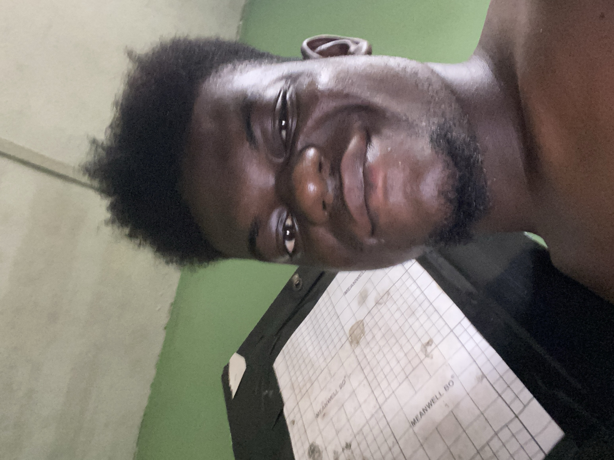 A close-up, low-angle portrait shows a Black man with dark, curly hair and a short beard. He is facing the camera with a slight smile. His skin appears to have a sheen, possibly due to moisture or lighting. A portion of his bare shoulder is visible.
In the foreground, there is a black clipboard with a white sheet of graph paper attached. The paper has smudges and text that appears to read "MEANWELL BO". The clipboard is positioned at an angle to the camera.
The background features a wall with a distinct line separating two colors: a light grey on top and a light green on the bottom. The specific location in the city of Unknown, Unknown is not discernible from the provided image.