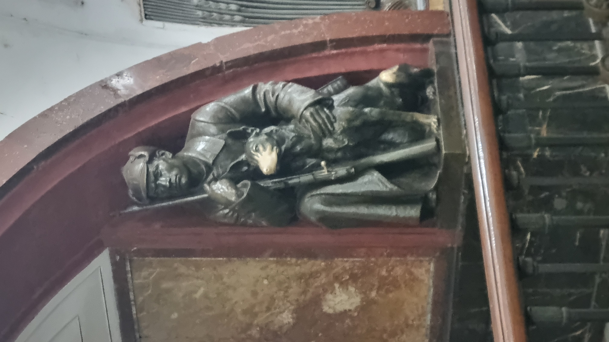 A bronze sculpture depicts a man in military attire, likely a soldier, kneeling beside a dog. The man wears a cap and a long coat, and holds a rifle vertically in his left hand, with the butt resting on the ground. His right arm is wrapped around the dog, which is also kneeling. The dog is medium-sized with its head turned to the left, looking slightly upwards. The sculpture is situated within an alcove featuring a dark red, curved architectural element above and a marbled-patterned panel to the left. In front of the alcove, a dark brown wooden railing with metal supports is visible. The overall setting suggests an indoor public space, possibly a metro station or a memorial.
