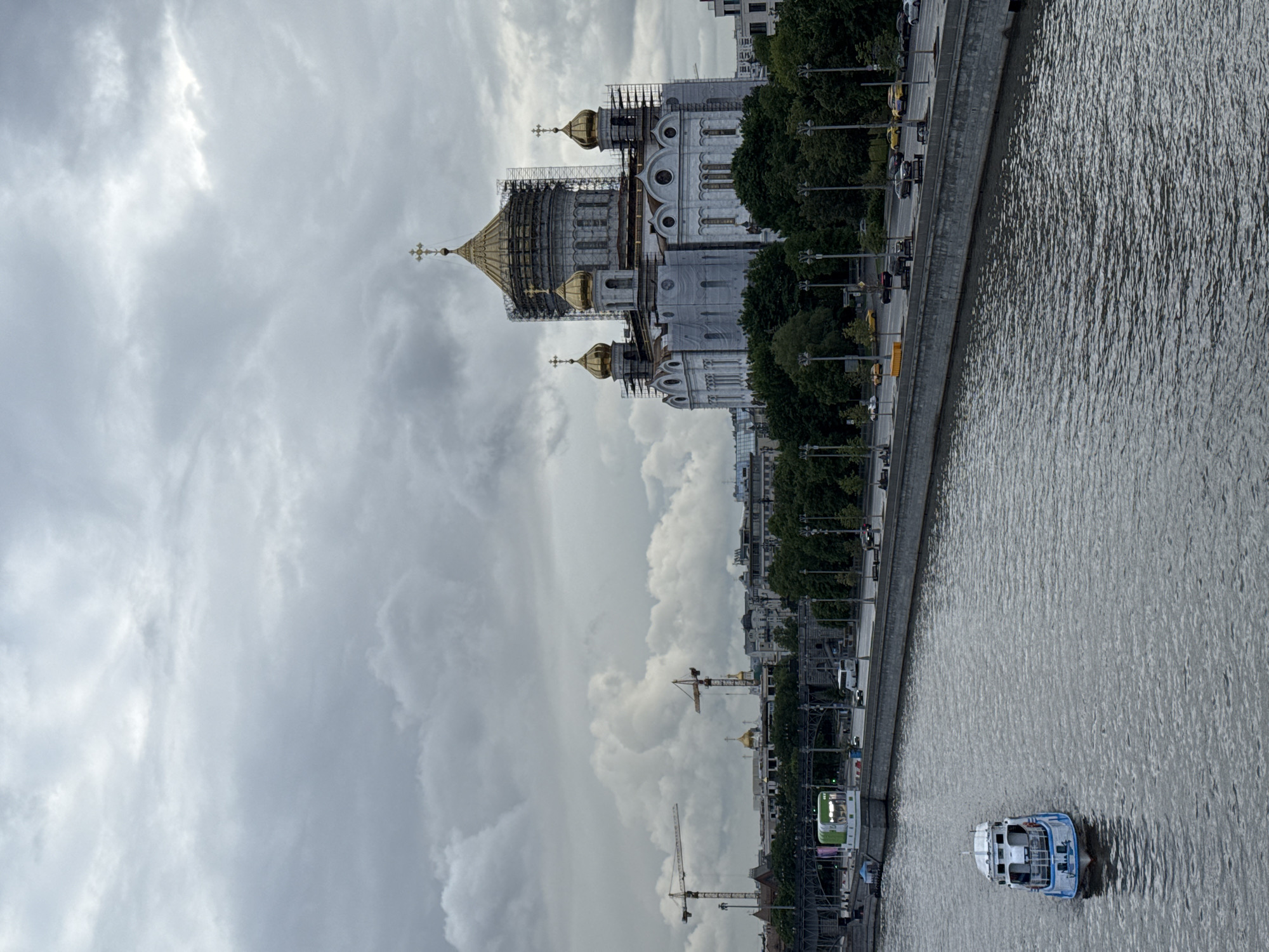 The image shows the Cathedral of Christ the Savior in Moscow, Russia, under a cloudy sky. The cathedral's golden domes are visible, with some parts appearing to be under renovation or repair, indicated by scaffolding. To the right of the cathedral is a river with a boat on it and a paved embankment lined with trees and streetlights. Cars and people can be seen along the embankment. In the background, construction cranes are visible among buildings. The overall scene conveys a sense of urban landscape juxtaposed with religious architecture and ongoing construction.