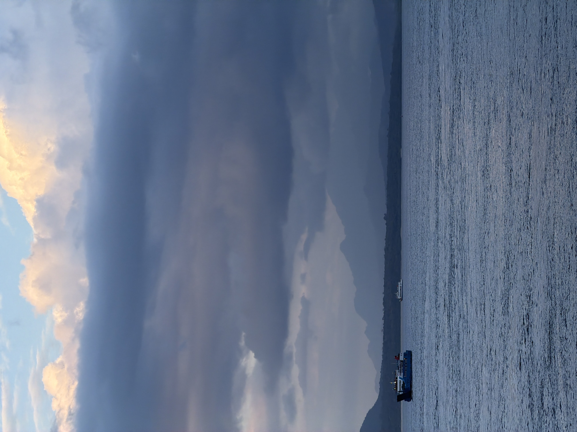 The image shows a body of water with two ferries in the distance, likely in the waters near Ketapang, Indonesia, given the context. The sky is filled with dramatic, dark clouds tinged with yellow and white where the sun is breaking through, suggesting either sunrise or sunset. The water is choppy and dark blue. In the foreground, the water fills most of the frame. Two ferries are visible in the middle ground. The ferry on the left is larger and blue, with lights illuminated on its deck. The ferry on the right is smaller and white, appearing further away. In the background, a hazy, dark mountain range lines the horizon, suggesting land is present beyond the water. The overall atmosphere is moody and serene.