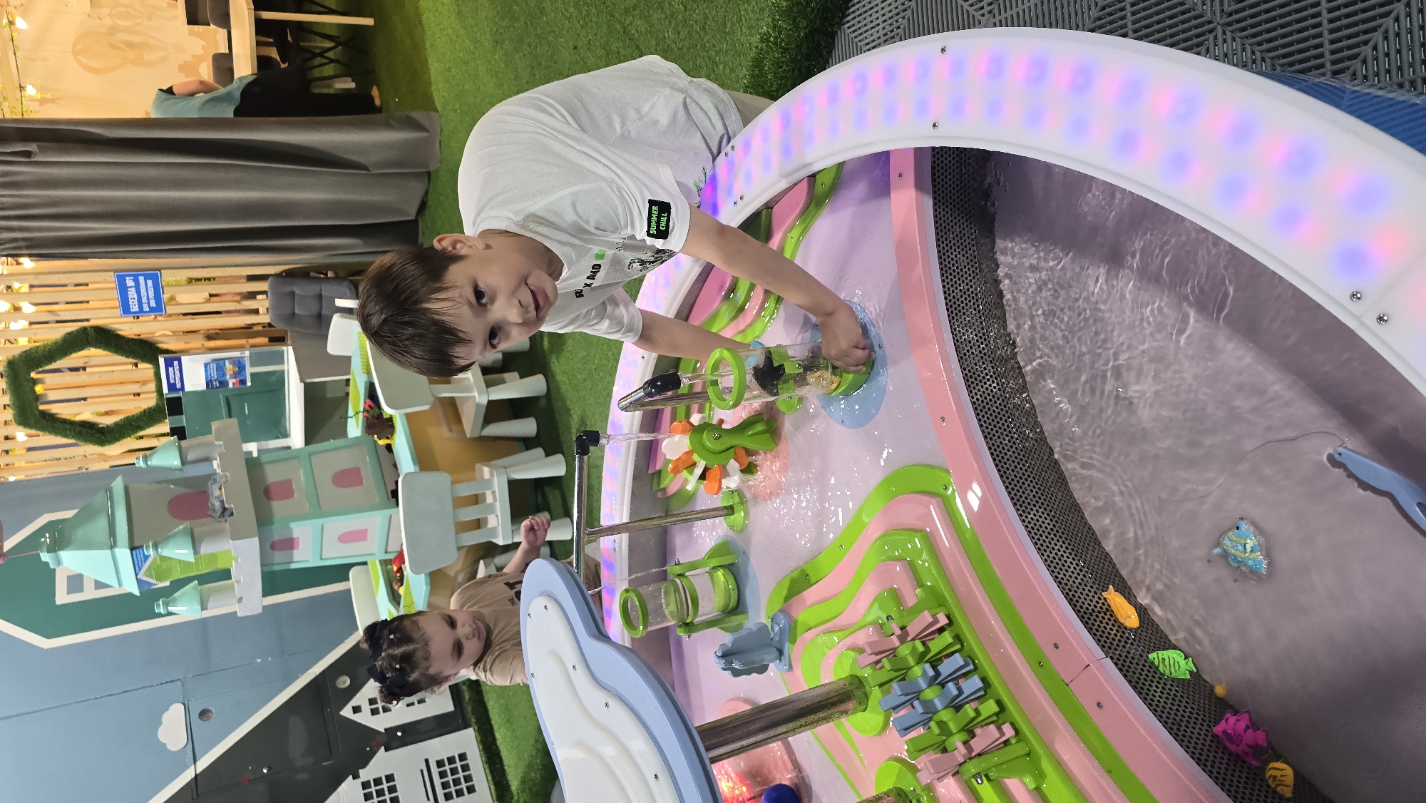The image depicts an indoor children's play area in Izhevsk, Russia, featuring a colorful water play table in the foreground. Two children are engaged in play. In the foreground, a boy, approximately 7-9 years old with short brown hair, stands leaning over a multi-level water table. He wears a white short-sleeved t-shirt with "SUMMER CHILL" text on the left sleeve, and beige shorts. His right hand is submerged in a clear, green-rimmed cylindrical container within the water table, manipulating its contents. Water flows from an overhead pipe into this container and other features. To the left of the boy, a girl, approximately 4-6 years old with dark hair styled in two pigtails, is also interacting with the water table. She wears a light brown t-shirt and looks down at the water. The water table itself is predominantly pink and light green, featuring various water flow elements. These include a large white and light blue "cloud" structure at one end from which water cascades, silver metal pipes directing water, transparent green cylinders, a multi-colored paddle wheel, and rows of small blue and pink paddle wheels along a wavy green track. The lower section of the table forms a shallow pool, which contains several small, brightly colored plastic fish toys (yellow, green, pink, blue) and a partially visible blue object resembling a fishing rod or net handle. The outer edge of the water table is illuminated by a strip of colored LED lights displaying pink, purple, and blue hues. A dark, perforated grate lines the inner edge of the lowest water level. The flooring directly around the water table is composed of grey interlocking soft tiles, transitioning to artificial green turf throughout the broader play area. In the mid-ground and background, various other play structures and environmental elements are visible. To the left, a light blue wall features white house outlines and a white cloud. Partially obscured behind the girl is a multi-level, light teal and white castle-like structure with pink accents. Small white children's chairs are scattered nearby. The background features a wooden slatted wall adorned with string lights and a large, green, hexagon-shaped decorative element made of artificial foliage. Signs written in Cyrillic, including "БЕСЕДКА №1" (Gazebo #1), are attached to this wall. A dark grey curtain partially screens a further section of the room, behind which a white table, chairs, and a partially visible individual are present. A dark
