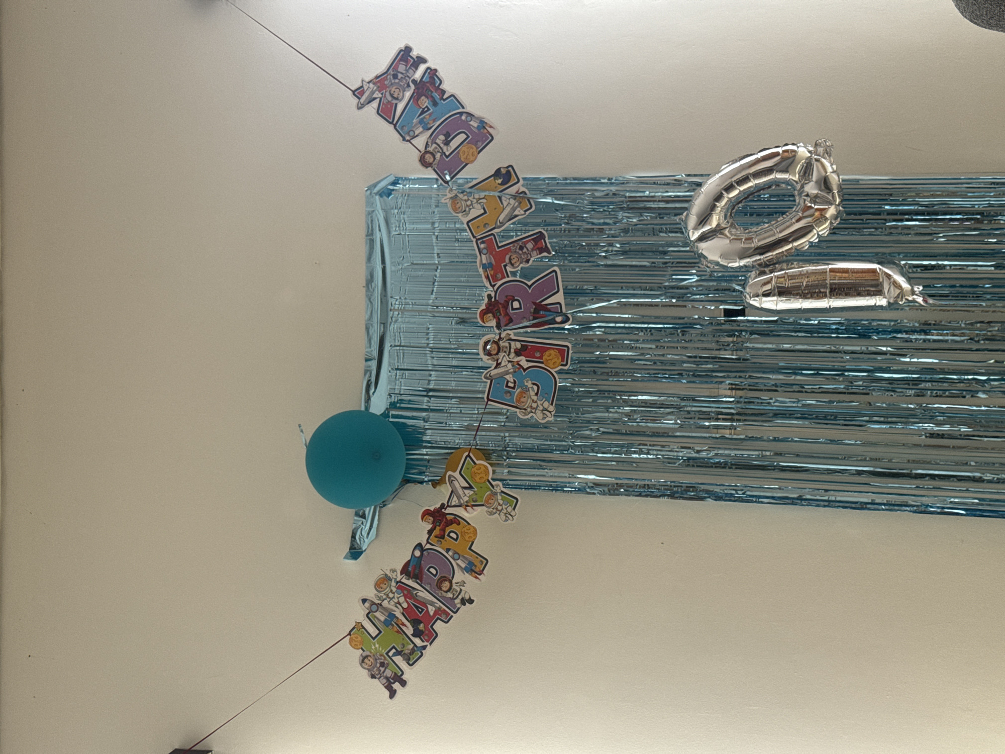 The image shows a birthday decoration hanging from a ceiling, likely indoors. The decoration includes a silver number "0" balloon and a long, silver sausage-shaped balloon. There's a blue tinsel background behind the balloons. Attached to the balloons are "HAPPY BIRTHDAY" banners with astronaut-themed designs. The scene also includes a single blue balloon.
The location is not identifiable from the image. No city or environmental details are visible to indicate it being in Kuta Selatan, Indonesia or anywhere else.