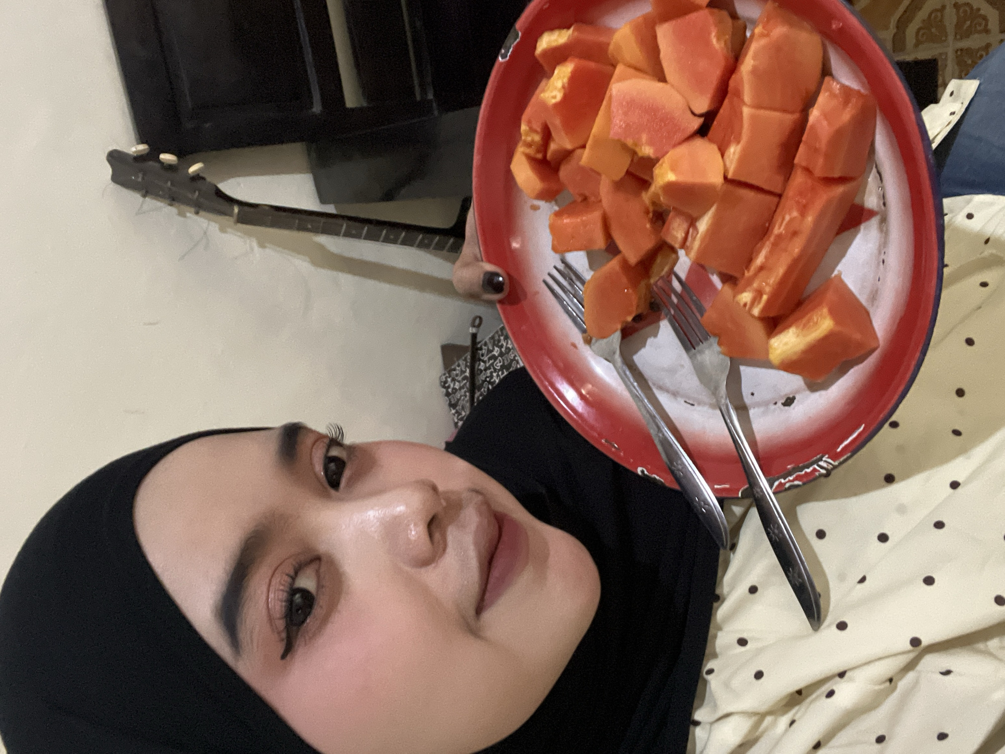 The image shows a close-up of a young woman wearing a black hijab and light-colored polka-dot attire. She is holding a red and white plate filled with chopped papaya. Two forks are visible on the plate. In the background, a dark-colored guitar leans against a wall, next to a dark piece of furniture. The scene appears to be indoors, possibly in a residential setting. No specific location in Bumi Mulyo, Indonesia can be determined from the visual information.