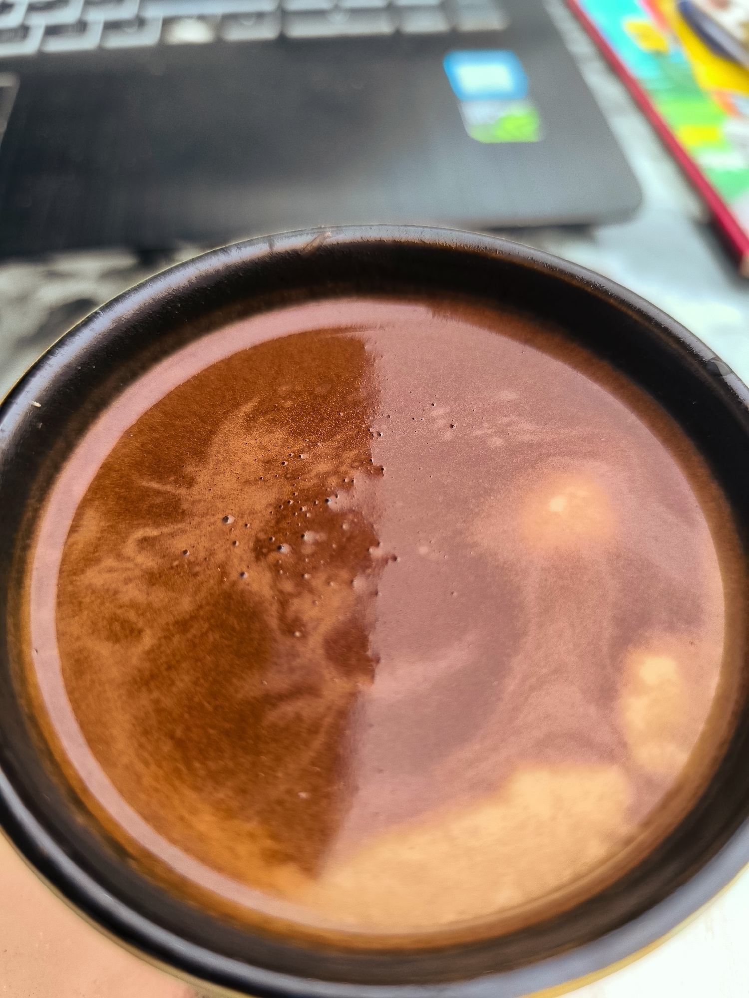 The image shows a close-up view of a dark liquid, likely coffee, in a black-rimmed mug. The surface of the liquid has a foamy texture with small bubbles and swirling patterns. In the blurred background, a laptop keyboard is visible, suggesting an indoor setting. The lighting creates reflections on the liquid's surface, highlighting its depth and color. There is no indication of the city of Batumi, Georgia, nor any specific location within it. The scene is static and focuses solely on the beverage and its immediate surroundings.