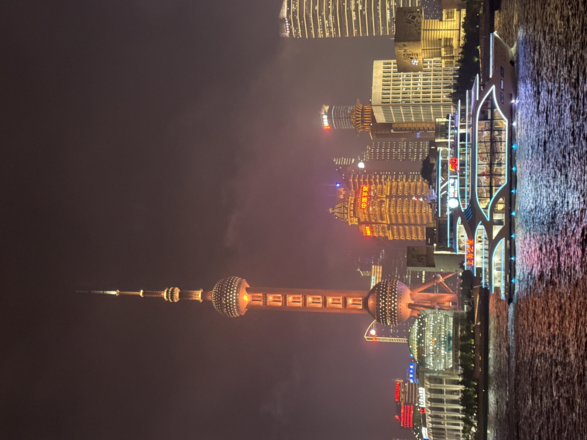 This media file captures a night view of the Shanghai skyline along the Huangpu River. The prominent subject is the Oriental Pearl Tower, a television tower in the Pudong district, illuminated with warm, orange lights. Other tall buildings, characteristic of a modern city, are visible in the background, their windows glowing with light. A boat with neon blue outlines is docked on the river, its reflection shimmering on the water's surface. The water appears choppy, with many small, bright reflections.
The scene is set at night, indicated by the dark sky and the artificial illumination of the city. The weather appears to be slightly hazy or misty, softening the lights and creating a dreamlike atmosphere. The location is clearly Shanghai, China, given the iconic Oriental Pearl Tower. No people are distinctly visible, and the focus is on the architecture and the urban landscape at night. The overall mood is one of urban vibrancy and nighttime spectacle.
There is visible text on some of the buildings, including Chinese characters. On the boat, some text is visible, including "滨江之光" (Bīnjiāng Zhī Guāng), which translates to "Riverside Light" or "Glory of the Riverside." Another sign on a building appears to have "MAG" and Chinese characters.