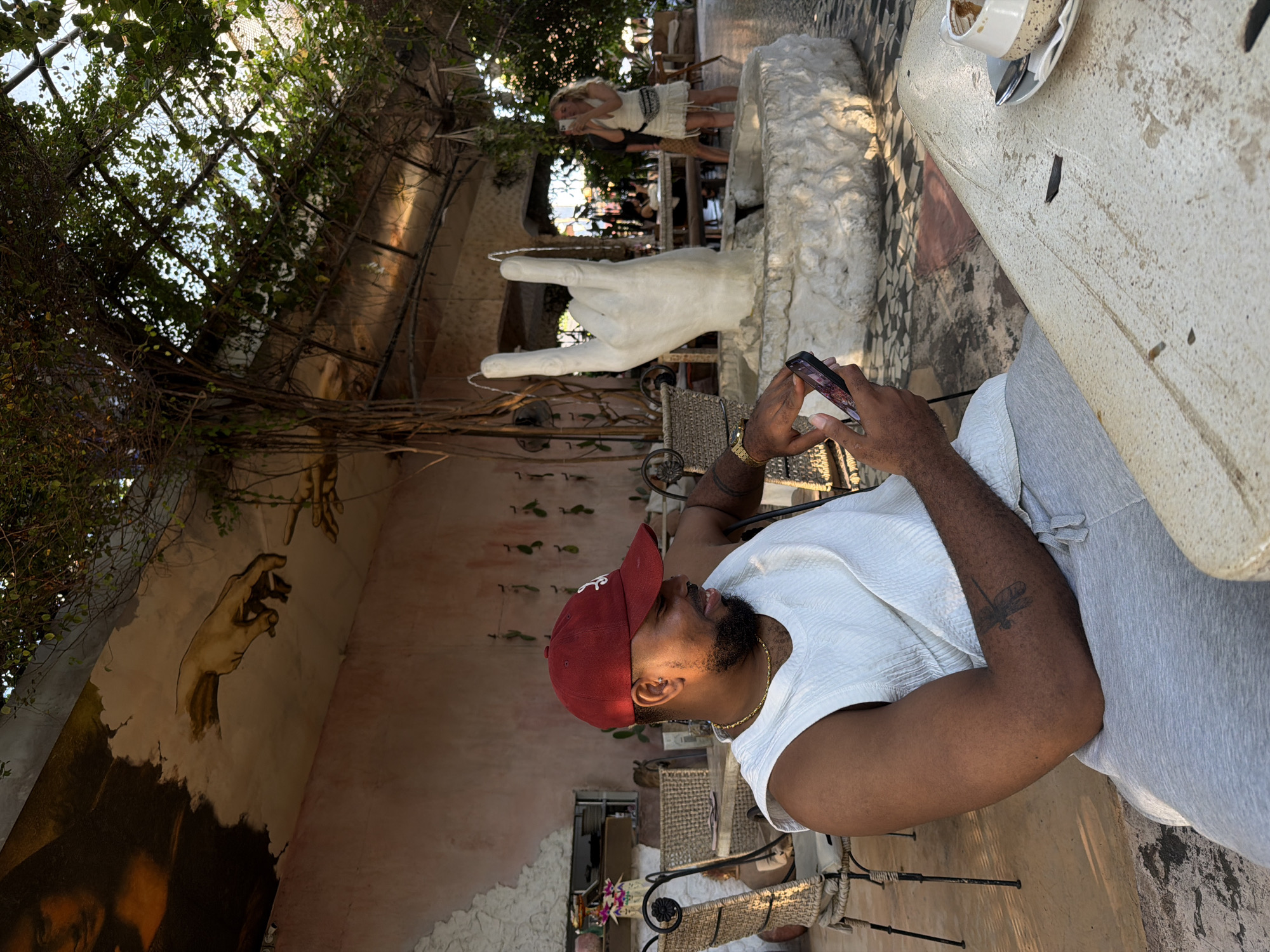 In North Kuta, Indonesia, a man wearing a red baseball cap and a white sleeveless shirt is seated at a table. He is looking down at his phone, which he holds in his right hand. A dragonfly tattoo is visible on his left arm. In the background, a large white hand sculpture serves as a fountain, with water spouting from its fingers. Behind the fountain, a woman in a white dress is taking a photo with her phone. The scene is set in an outdoor seating area with tables and chairs, surrounded by greenery and an overhead structure covered in vines. A mural depicting hands reaching out is visible on the wall in the background.