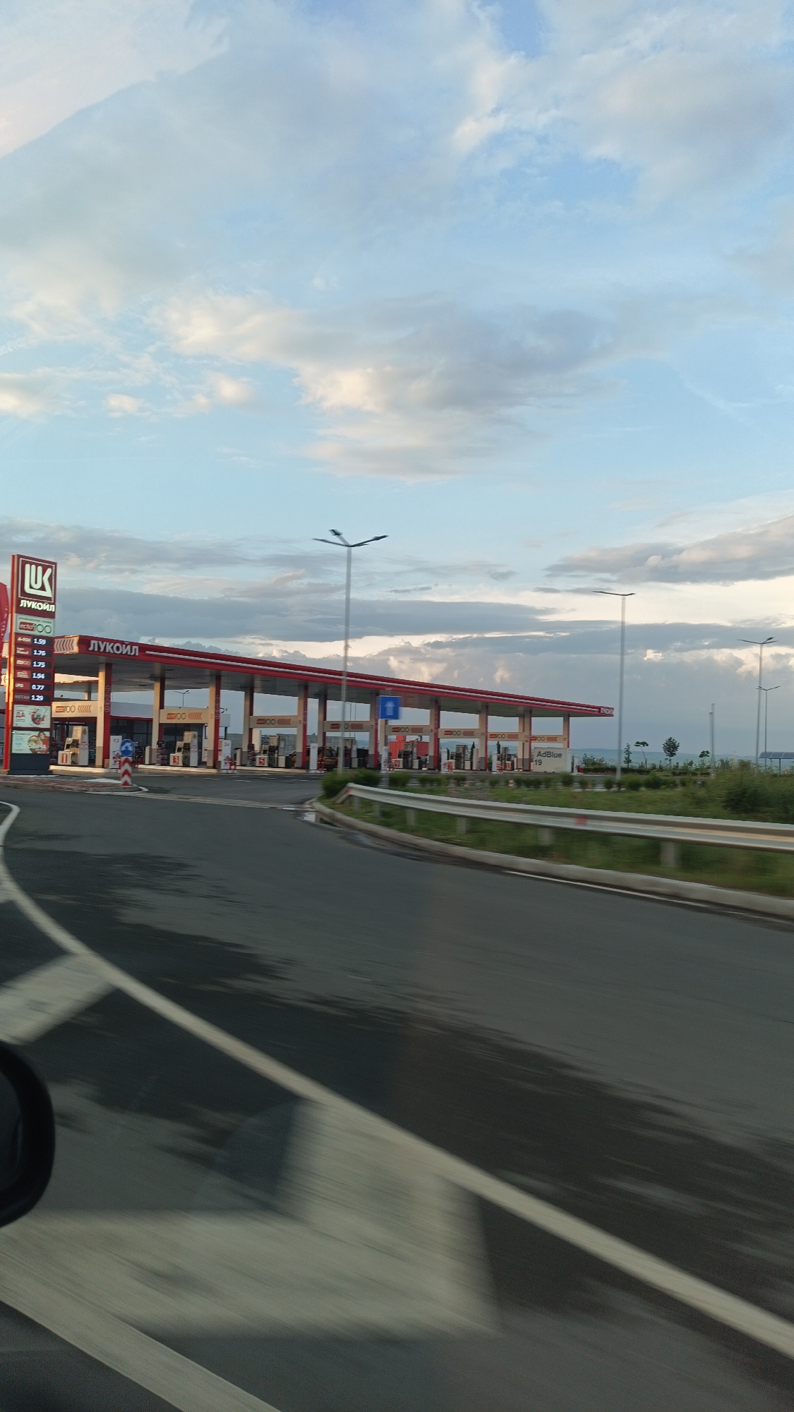 The image shows a Lukoil gas station located in Burgas, Bulgaria. The station has a red and white color scheme, with a sign displaying fuel prices. The station has multiple gas pumps under a large canopy. A white guardrail is visible along the roadside. The road surface is wet and reflects the sky, indicating recent rain. The sky is partly cloudy with a mix of blue and gray hues. The photo appears to be taken from a moving vehicle.