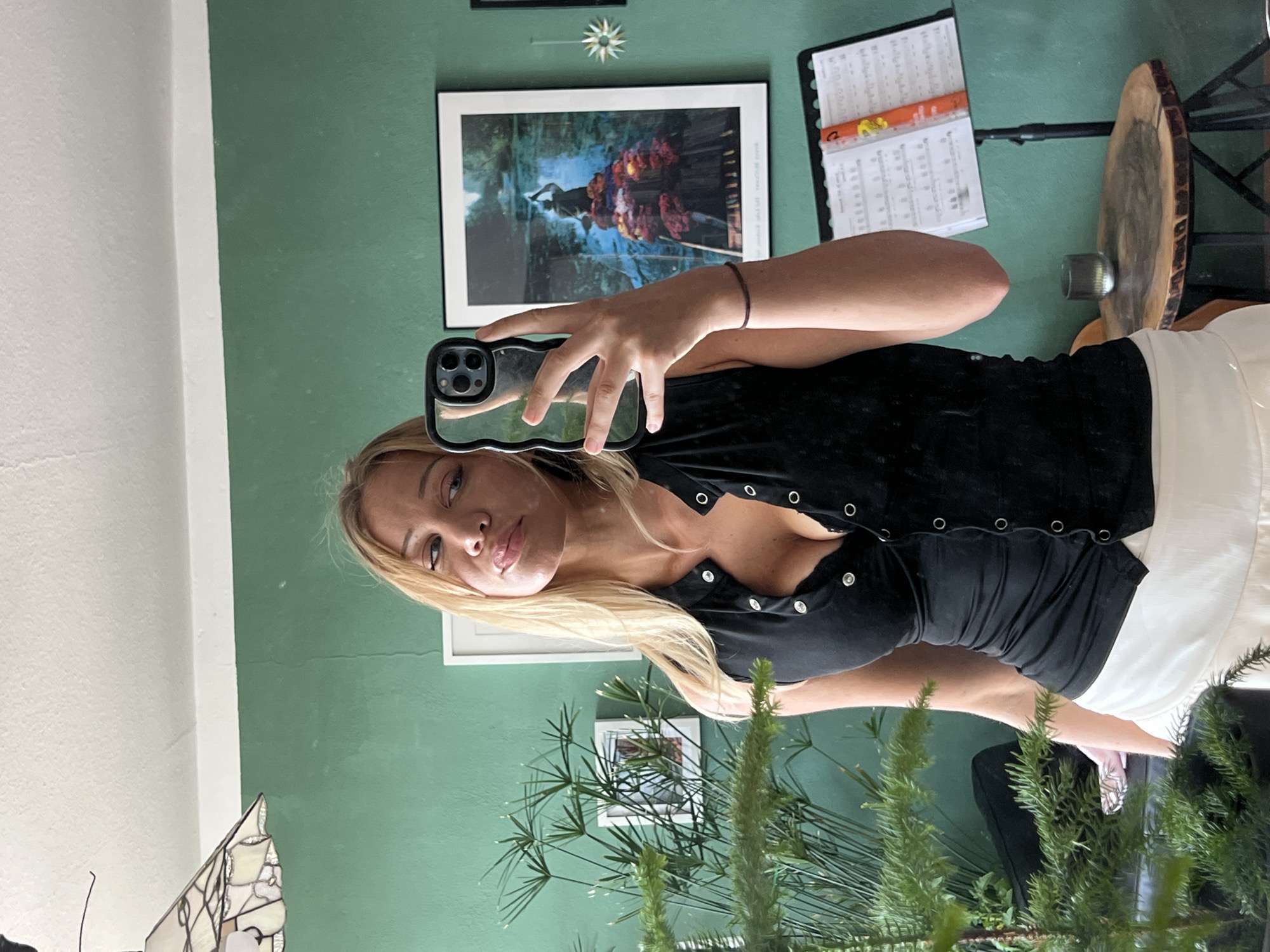This is a selfie taken by a young woman with blonde hair and blue eyes. She is wearing a black, button-up top and a white skirt. Her expression is relaxed and she is looking directly at the camera. The background is a green wall with several framed pictures and a music stand with sheet music. A large potted plant with spiky green leaves is in the foreground. The image is oriented vertically, suggesting it was taken with a phone. The lighting appears to be natural, possibly from a window, and the time of day is difficult to determine. The setting appears to be indoors, likely a home or studio. There is no visible text in the image. The location context provided is Mühldorf a. Inn, Germany.