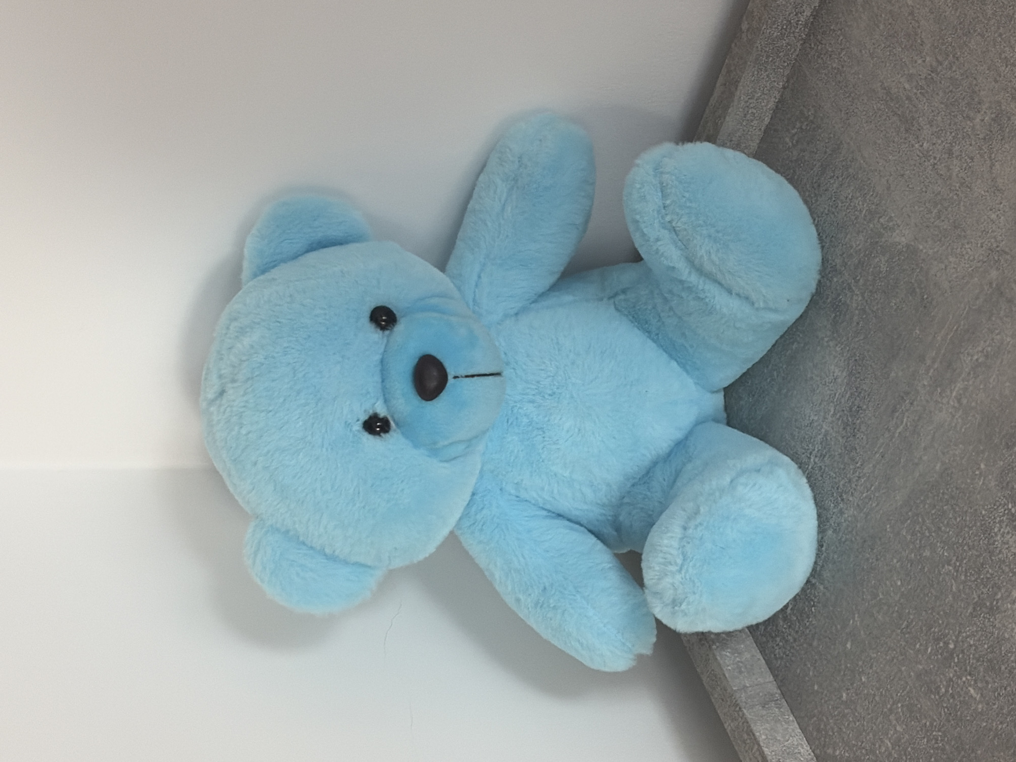 A light blue plush teddy bear sits on a grey, textured surface. The bear is facing the viewer, with its paws resting on the surface in front of it. Its eyes are black, and it has a small, dark brown nose and a stitched mouth. The bear is positioned in the corner of a room where two white walls meet. The lighting suggests an indoor environment. There is no indication of the city of Hanniv, Ukraine within the image.