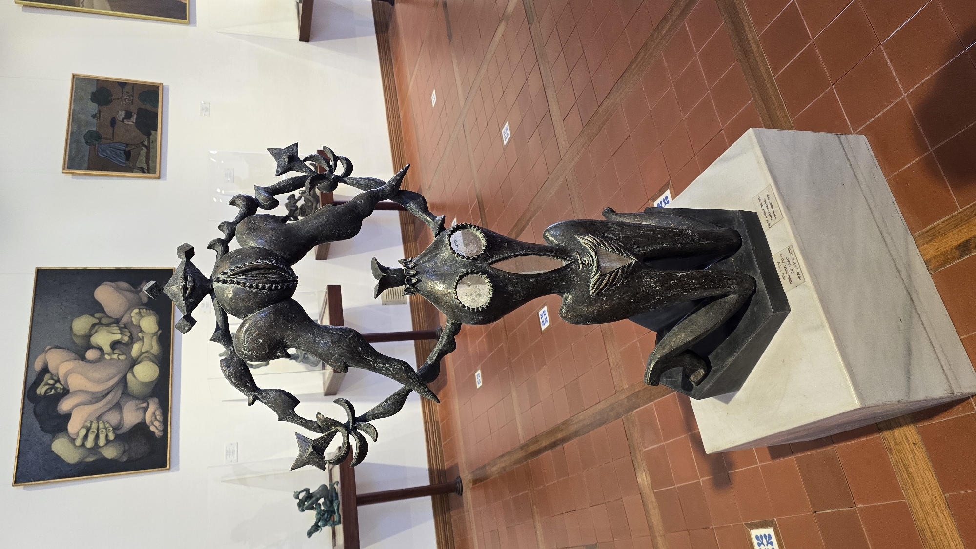The image shows a bronze sculpture of a stylized, humanoid figure with an abstract, almost monstrous, headpiece, and outstretched arms ending in star-like forms. The figure is kneeling on a marble base, which is placed on a terracotta tiled floor. Behind the sculpture, on a white wall, are two paintings. The painting on the left is abstract and depicts intertwined figures in muted colors. The painting on the right is a landscape with trees and figures. The overall setting appears to be an art gallery or museum. The base of the sculpture has a plaque with text in Hebrew and English, indicating it is part of an exhibition. The location is identified as the City of Hof HaCarmel Regional Council, Israel, suggesting this is a public art space within that region.