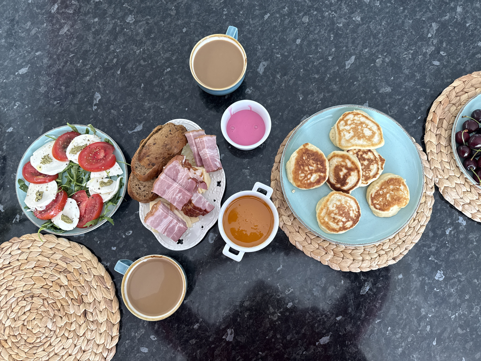 The image displays a top-down view of a breakfast spread on a dark, speckled countertop. To the upper left, a round, woven placemat sits next to a light blue mug filled with coffee. To the right of the placemat, a light blue plate holds a Caprese salad consisting of sliced tomatoes, mozzarella, and arugula, drizzled with a green sauce. Below the coffee mug, a white dish contains several strips of bacon and slices of bread. Further down, a white bowl with handles holds a reddish-orange sauce, and next to it, a small white dish contains a bright pink liquid. In the lower half of the image, a light blue plate with a woven placemat underneath features a stack of small, golden-brown pancakes. Another woven placemat is visible in the bottom right corner, partially showing a light blue dish filled with dark red cherries. There are no people or animals present in the image. The scene is set indoors, likely in a kitchen or dining area. There is no information within the image that indicates the location is the city of Sochi, Russia.