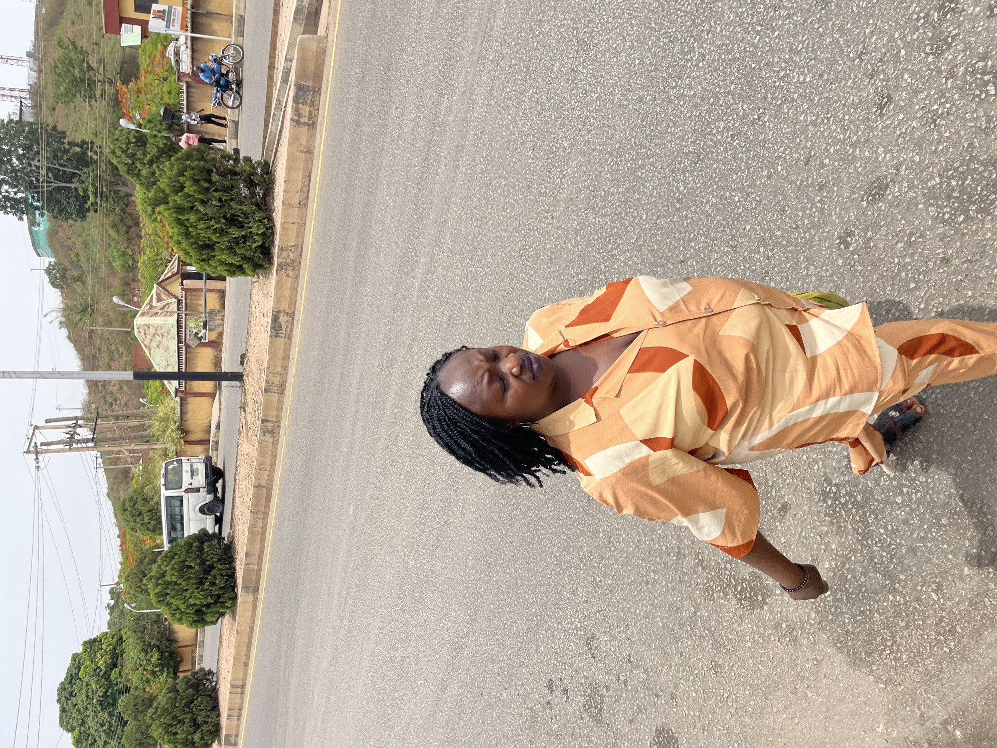 The image displays an outdoor street scene in Jos, Nigeria, during what appears to be a bright daytime. The primary subject is a dark-skinned individual with braided hair, dressed in a patterned orange, brown, and cream two-piece outfit (shirt and trousers), and dark sandals. They are standing on a paved road, their eyes seemingly closed or cast downwards, giving a calm or contemplative expression.
The road, made of light grey asphalt, occupies a large portion of the frame, featuring a yellow line marking its edge. In the background, a verdant landscape unfolds, with various green trees and shrubs lining the roadside and covering a gently sloping hill. Further along the road, a white mini-van is parked on the shoulder, and utility poles with multiple wires are visible.
Beyond the parked vehicle, another section of the road continues, bordered by more vegetation and small buildings, including one with a green roof and another with a reddish-orange roof. On the opposite side of the road from the main subject, a blue motorcycle with two riders, along with at least two other individuals standing nearby, can be seen. A sign on a building in this area is partially visible, reading "NATIONAL COLLEGE OF...". The lighting suggests a clear day, possibly with some atmospheric haze.