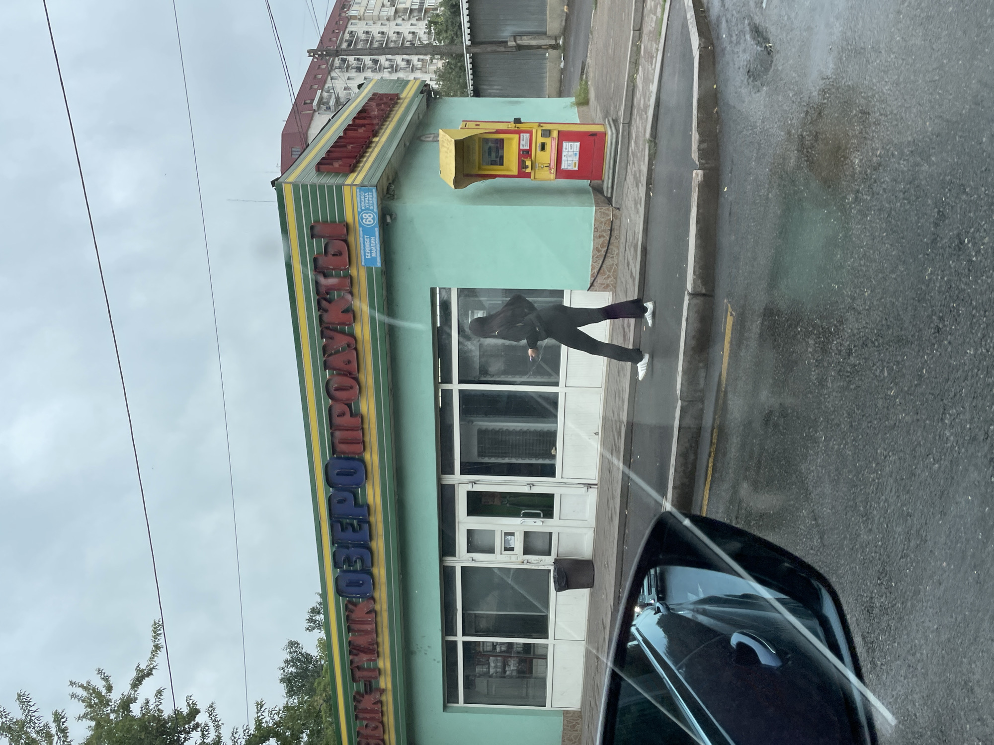The image shows a street view in Almaty, Kazakhstan, under an overcast sky. In the foreground, a car's side mirror reflects the street and other vehicles. The main subject is a pale green storefront with a yellow and green striped awning. Large, bold letters on the awning spell out "ОЗЕРО ПРОДУКТЫ" (Lake Products) in blue and red. A blue street sign above the entrance reads "БЕЙМБЕТ МАЙЛИН" and includes the number "68" within a circle, along with "УЛИЦА STREET".
A lone individual wearing a black hooded jacket and dark pants with white stripes down the sides walks along the sidewalk in front of the store, holding a smartphone. To the right of the entrance, a red and yellow ATM machine stands next to the building. The sidewalk is wet, suggesting recent rain, and puddles are visible on the asphalt. Behind the storefront, an apartment building with multiple balconies is visible. A metal fence and some trees complete the background. A trash can is placed near the store's entrance.