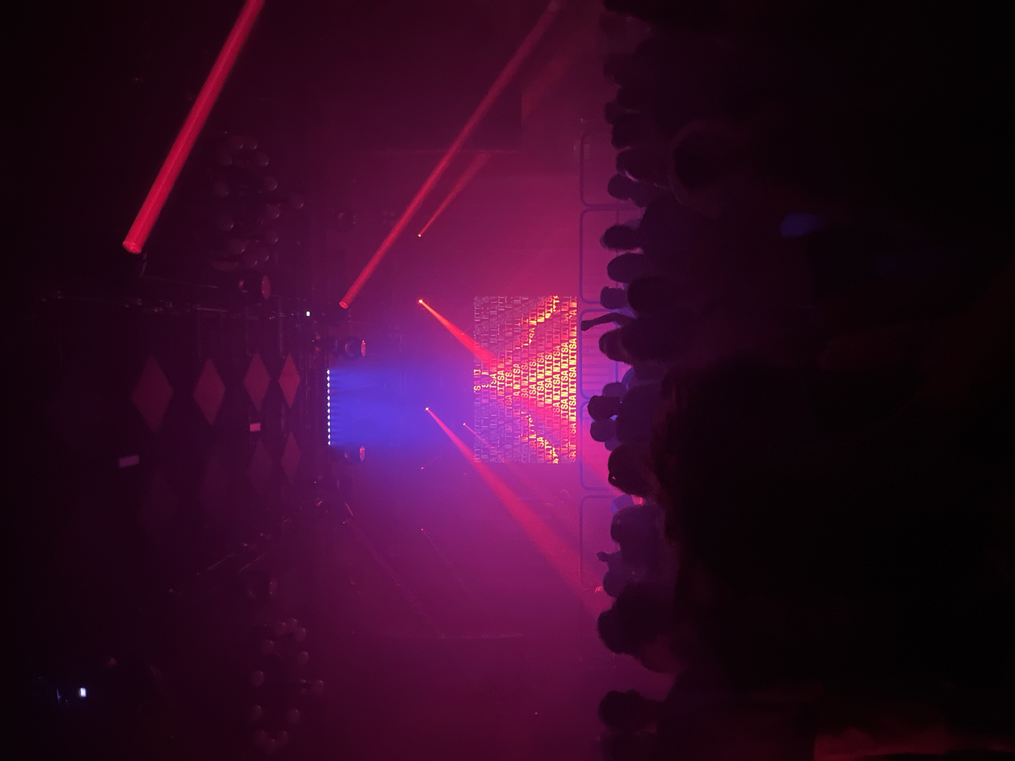 The image depicts a crowd of people at a dimly lit indoor event, likely a concert or club. Red and blue lights illuminate the scene, with red beams cutting across the frame from above. In the background, a screen displays repeated text in a light color against a darker background. The crowd is largely silhouetted, with their forms blurred by the low lighting. The environment appears to be a venue with a dark ceiling and some decorative elements, possibly diamond-shaped, visible at the top. The image suggests a lively atmosphere with a focus on the light show and the performance. The location of this event is Pacha Barcelona, a well-known nightclub located at Port Olímpic, 20, 08003 Barcelona, Spain.