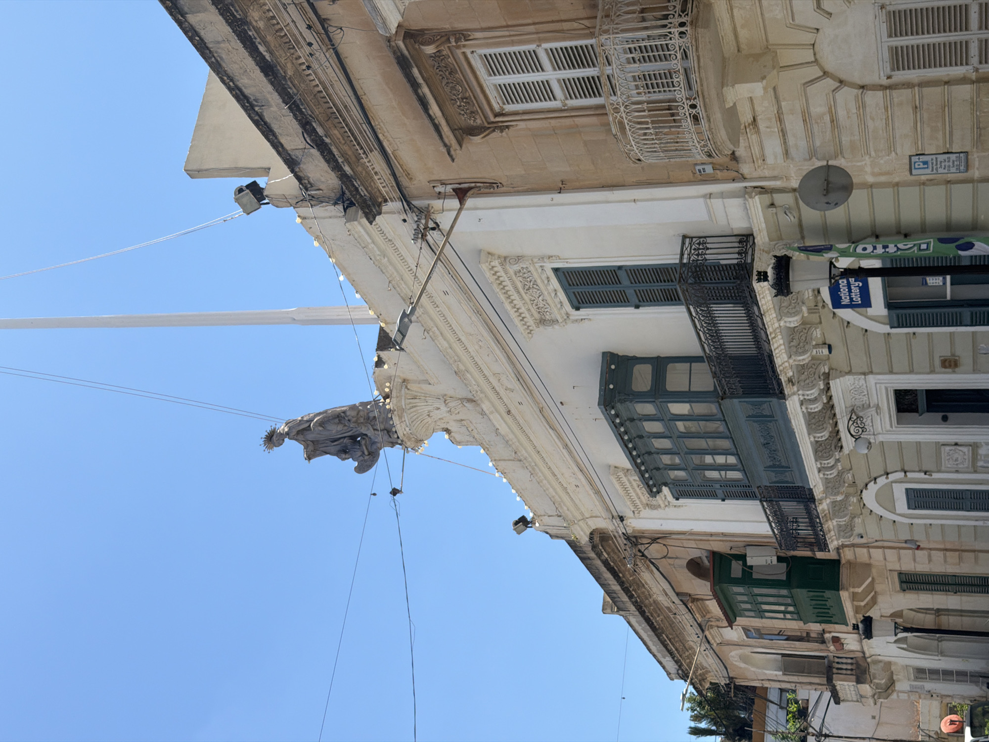 The image shows a street view in Sliema, Malta. The scene is dominated by the facades of old buildings with balconies and ornate architectural details. A statue of what appears to be a religious figure with a halo stands atop one of the buildings, silhouetted against a clear blue sky. There are power lines and a tall flagpole visible.
One building features a dark green enclosed balcony with multiple panes. Adjacent to it, another building has a white wrought-iron balcony. Several windows with closed shutters are visible on the different structures. A lamppost with a street light is positioned in the foreground. A sign for "National Lottery" is attached to the lamppost. The buildings are constructed with a mix of stone and plaster, in shades of beige, cream, and white. The overall impression is one of a sunny day in a historic European city.