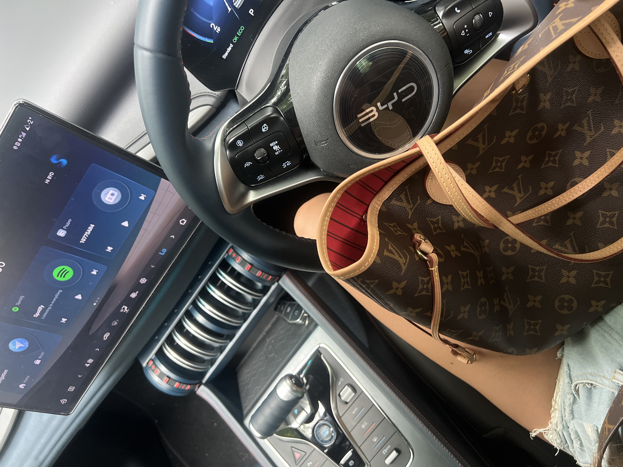 The image shows the interior of a car, likely a BYD, with a focus on the central console and steering wheel. A person's legs are visible, wearing denim shorts, with a Louis Vuitton handbag placed across their lap. The car's touchscreen display shows navigation and Spotify menus. The steering wheel has control buttons, and the gear shifter is visible on the console. The environment appears to be a car interior, with no clear indication of an outdoor urban setting.