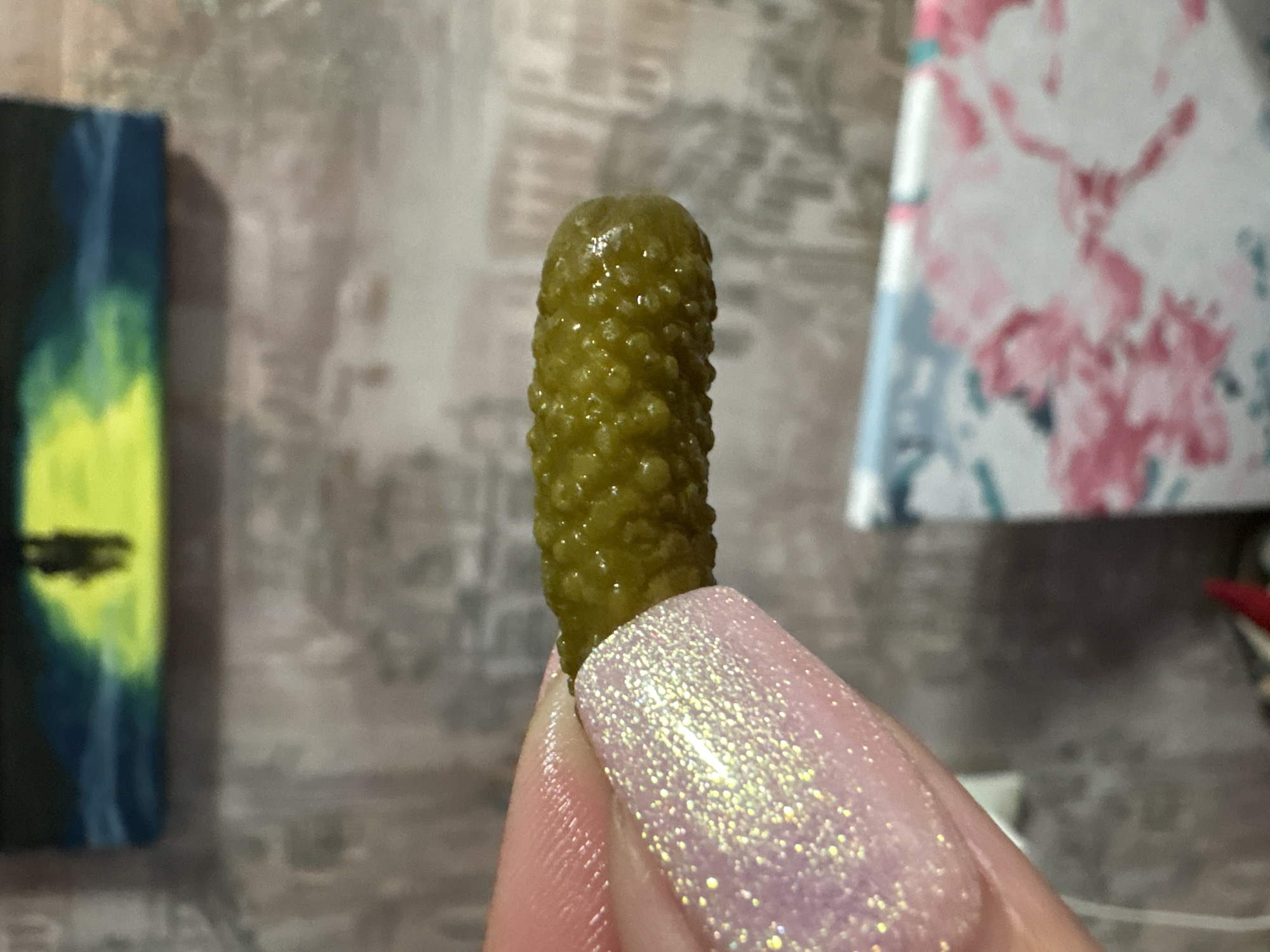 The image features a single green gherkin pickle being held by a person. The pickle is in sharp focus, exhibiting its characteristic bumpy texture and glossy surface. The person's fingers are visible, adorned with long, light pink nails covered in glitter polish. The background is blurred, showing two paintings, a grey tiled wall and what appear to be items on a desk. The image suggests a close-up shot indoors with a focus on the pickle and the person's hand. There is no information to determine the location in the city of Марковский, Russia.