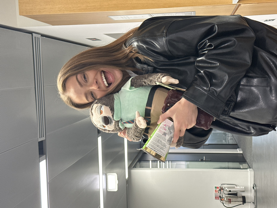 A female subject, with light brown hair and a broad, open-mouthed smile, is centered on the right side of an indoor hallway. She wears a black faux-leather jacket. In her arms, she cradles a plush toy resembling the character Flash the sloth from Disney's Zootopia, which is dressed in a light green shirt and a striped tie. Her left hand also holds a packaged food item with green and white labeling, positioned above a dark red smartphone encased in a textured cover. The background consists of a modern interior corridor featuring white perforated acoustic ceiling tiles with integrated rectangular LED light panels. A white wall is visible on the left, with a dark-framed glass door further down the hallway. A fire extinguisher station, comprising two red extinguishers on a chrome stand, is present on the far left. A light wooden panel wall is partially visible on the right. The flooring is a neutral grey. The specific location within London, United Kingdom, cannot be definitively identified from the provided visual information, but the environment is consistent with a contemporary office building or institutional facility.