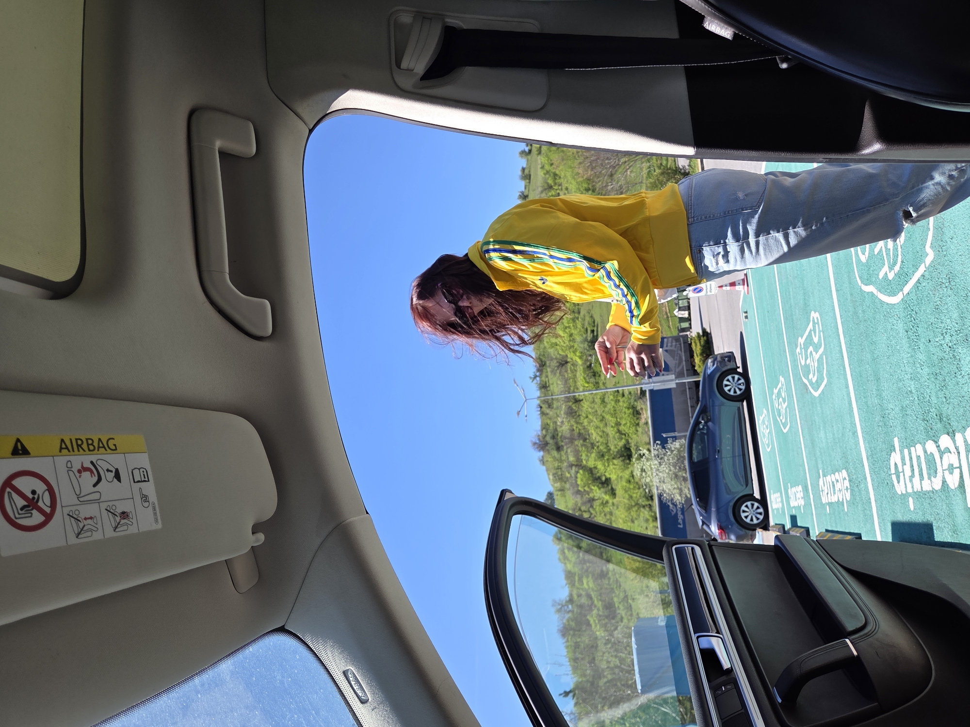 The view is from inside a car, looking out the open driver's side door at a parking lot. A person wearing a yellow jacket with blue stripes and jeans stands in the parking lot. Another car is parked next to the open door. The parking lot has green painted parking spaces with white logos and text. Trees and a hill are visible in the background. The sun visor is down on the driver's side, with an "AIRBAG" warning label visible. Based on the "e-lectri.fi" text in the parking lot, the location is Levunovo, Bulgaria.