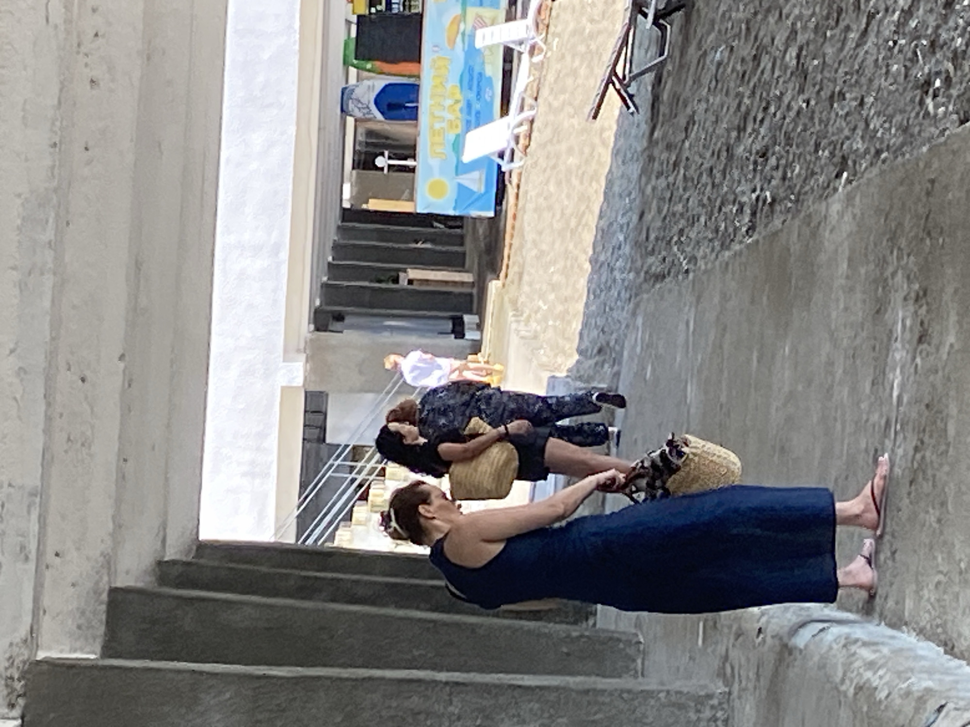 The image shows a beachside scene in Chabkwara, Abkhazia, under a concrete structure. In the foreground, a woman wearing a dark blue dress and flip-flops stands facing away from the camera, holding a straw bag with a patterned fabric insert. To her right, another woman in a black top and shorts carries a similar straw bag. Further back, a person in a camouflage uniform and another individual in a white shirt and shorts are visible walking. Behind them, a banner with "ЛЕТНИЙ БАР" (Summer Bar) written in Cyrillic is displayed, along with deck chairs and surfboards. The ground is a mixture of sand and concrete.