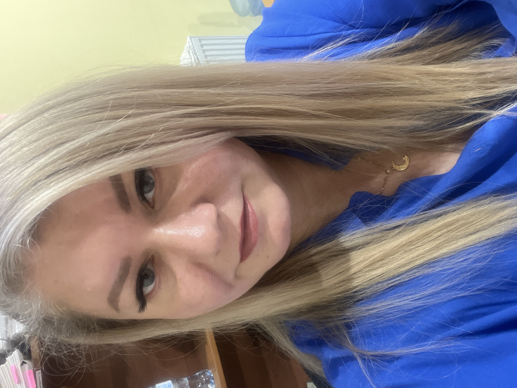 A woman with light blonde hair and fair skin is shown in a close-up selfie-style photograph. She is wearing a bright blue top, and a gold necklace with a crescent moon charm is visible around her neck. Her makeup includes dark mascara or false eyelashes. The background is blurred, but a yellowish wall and part of what appears to be a white radiator are visible. The image doesn't include any information to conclude the location in the city of Izhevsk, Russia.