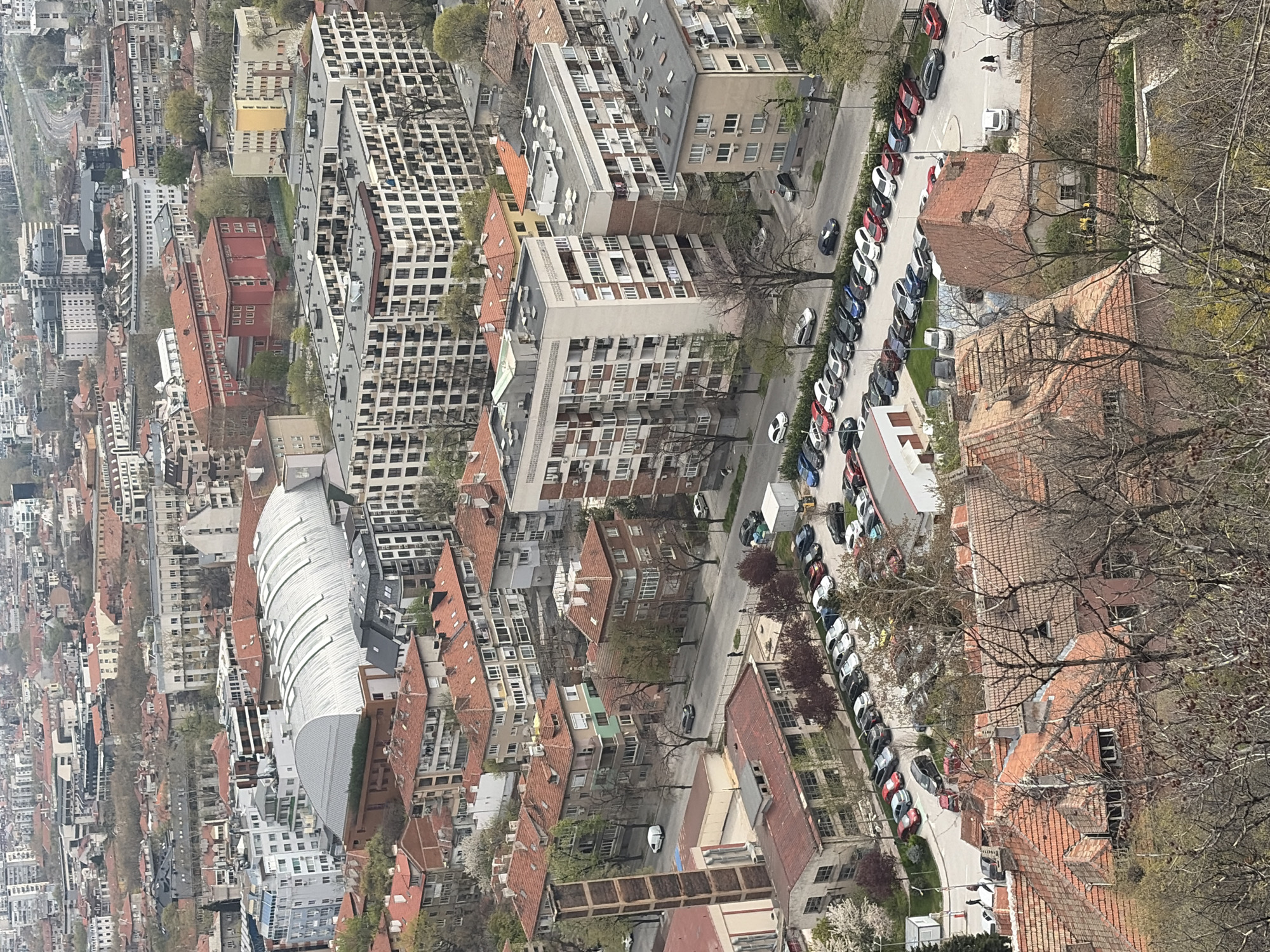 The image is an aerial view of a section of Plovdiv, Bulgaria, taken on a clear day. The scene is dominated by dense urban development, featuring buildings with red-tiled roofs and modern structures.
A long row of parked cars lines the street, with a mix of colors including white, red, black, and blue. Pedestrians can be seen walking along the sidewalk near the parked cars. The building architecture is diverse, ranging from older structures with pitched roofs to larger apartment blocks with multiple stories.
There are signs of green spaces visible between the buildings, with trees interspersed throughout the cityscape. A curved building with a light-colored roof stands out amongst the other structures. Bare tree branches are visible in the lower right corner of the image.