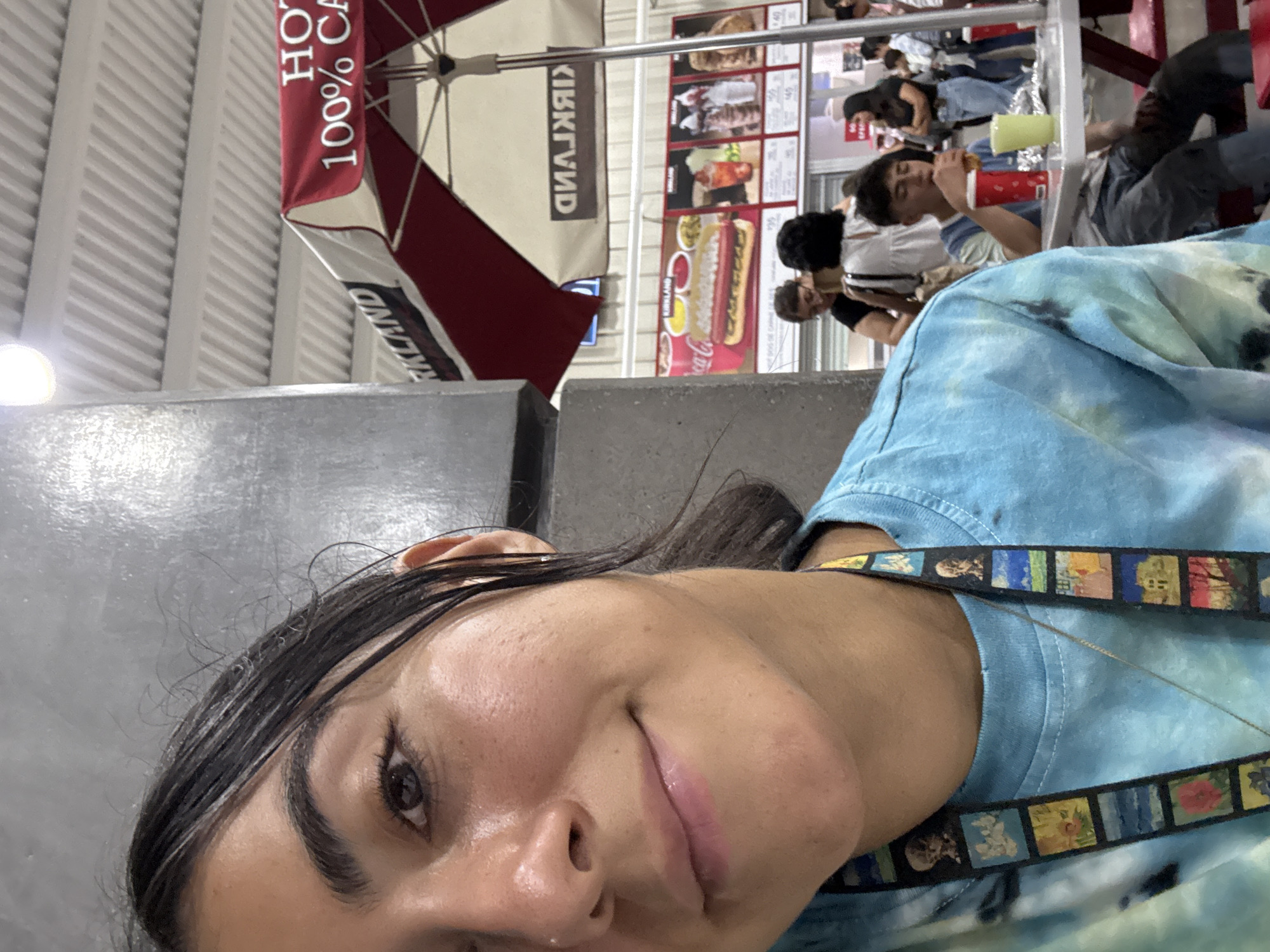 A selfie shot shows a woman with dark hair, fair skin, and a blue tie-dye shirt with a lanyard around her neck. Behind her is a Costco food court setting. Patrons sit at tables, some eating. A menu board displaying food options and pricing is visible. A large red and white umbrella bearing the "Kirkland 100% CA" text shields customers. This setting suggests the photograph was taken at a Costco location. Given the prompt's explicit location, it is highly likely this image was taken at the Costco food court in the city of Puebla, Mexico.