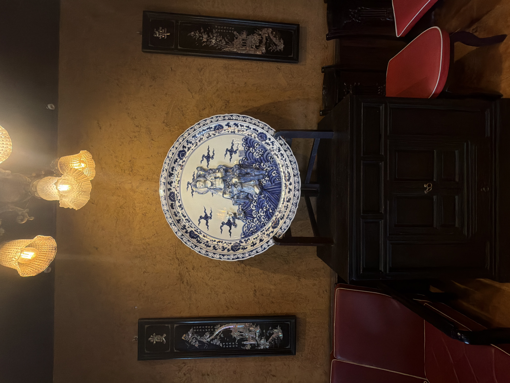 This image captures a dimly lit indoor scene with traditional East Asian decor. The focal point is a large, ornate blue and white porcelain plate depicting figures and waves, displayed on a dark wooden stand atop a dark wooden cabinet. Flanking the plate on the wall are two tall, narrow black panels with intricate inlaid designs, likely mother-of-pearl. Above, three woven light fixtures with visible bulbs cast a warm glow. To the left, a section of a red upholstered sofa with white trim is visible. To the right of the cabinet, dark wooden chairs with red seats are partially visible, suggesting a dining or seating area. The wall has a textured, yellowish-brown finish. The overall ambiance suggests an interior space, possibly a restaurant or a home, decorated with Asian cultural artifacts. Without additional context, the specific location in Đà Nẵng, Vietnam cannot be determined.