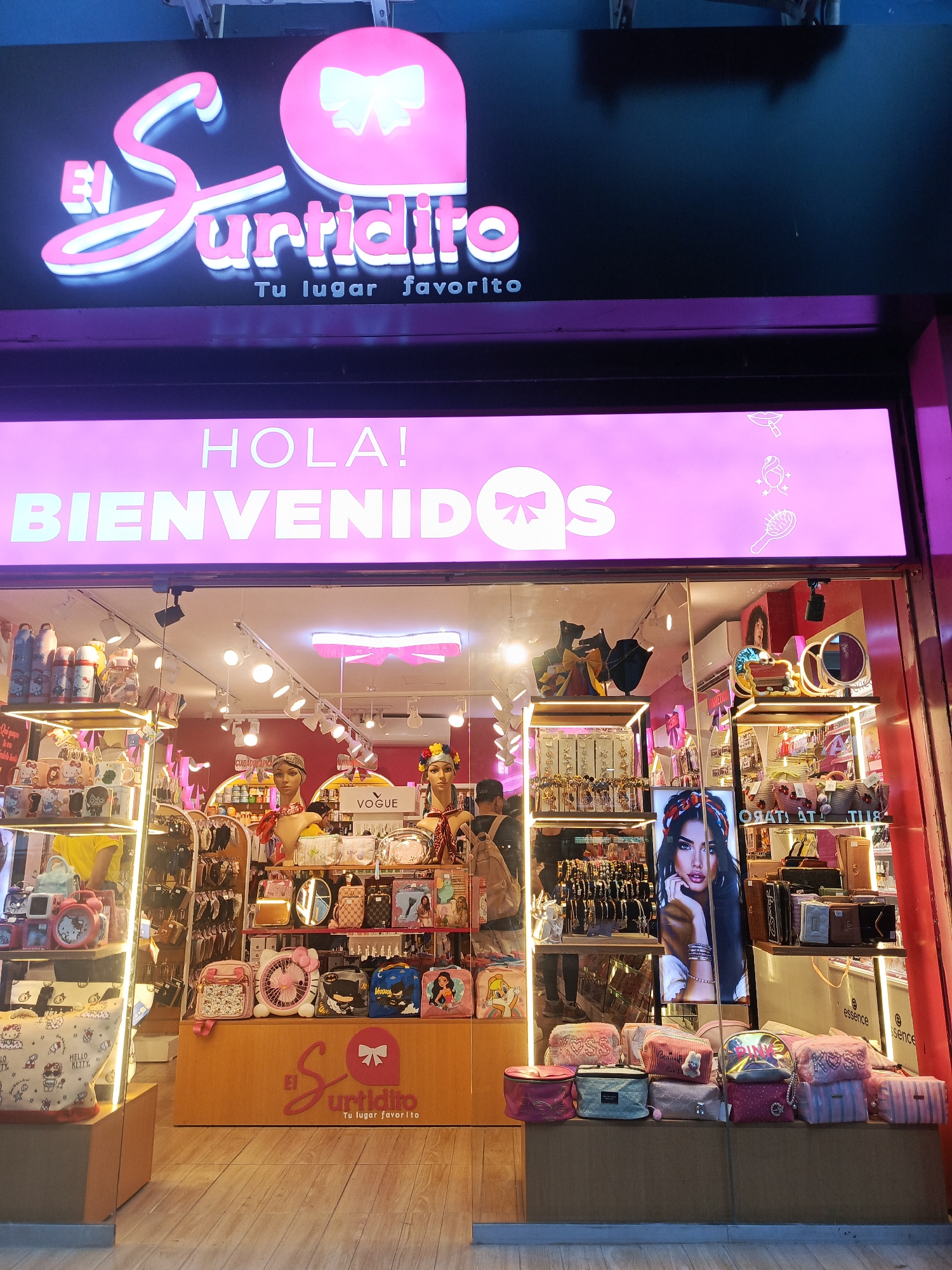 The image shows the storefront of "El Surtidito," a shop located in Guayaquil, Ecuador. The storefront features a bright pink neon sign with the shop's name and logo, which includes a stylized bow. Below the main sign, a banner with "HOLA! BIENVENIDOS" is illuminated in white. The interior of the shop is visible through a large glass window, displaying shelves filled with various merchandise such as bags, cosmetics, and decorative items. Mannequins are positioned in front of the shelves, showcasing some of the products. The shop appears to be well-lit and stocked, suggesting it is open for business. The flooring in front of the store is made of wooden planks.