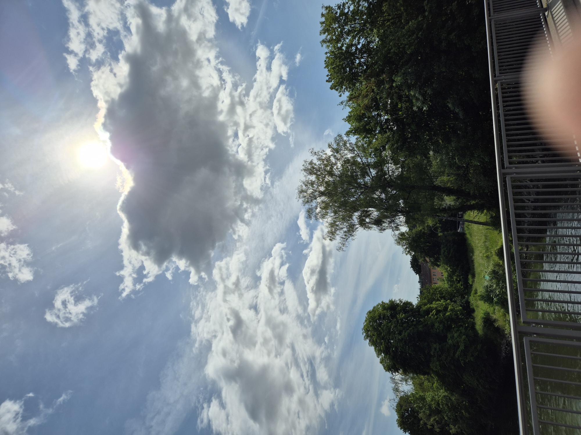 The image presents an outdoor, daytime scene under a partly cloudy sky in the city of Unknown, Unknown. The upper half of the frame features a bright sun in the upper-center, partially obscured by a large, dense grey cloud, creating a distinct sunburst and lens flare. Numerous white cumulus clouds are scattered across the clear blue sky. The middle ground is dominated by a dense line of green deciduous trees and bushes. A prominent light green tree stands near the center, while darker green trees flank it. In the background, behind the trees on the right side, a light-colored, possibly pinkish-orange, multi-story building with a dark roof is visible, along with a patch of green grass. The lower portion of the image shows a dark green body of water, likely a river or canal, beneath a grey metal bridge railing that extends horizontally across the foreground. A blurry, light-skinned human finger or thumb is visible in the bottom right corner of the frame, partially obscuring the bridge railing and water.