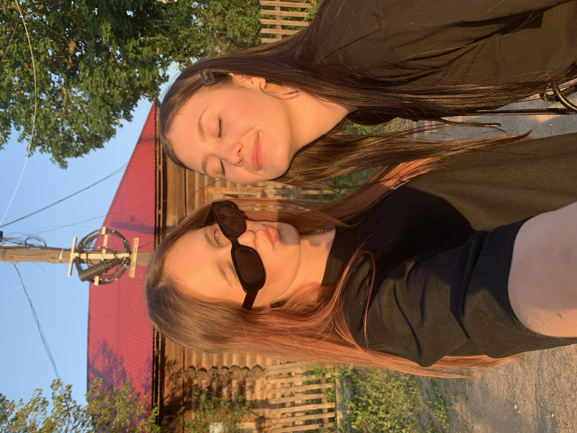 Two young women are pictured outdoors. One woman with long dark hair is lying behind the other, her eyes closed. The second woman, with long reddish-brown hair, wears sunglasses. Both women are wearing black shirts. Behind them is a red-roofed wooden building. A power pole with wires is visible on the left side of the image. The location is Фоки, Russia.