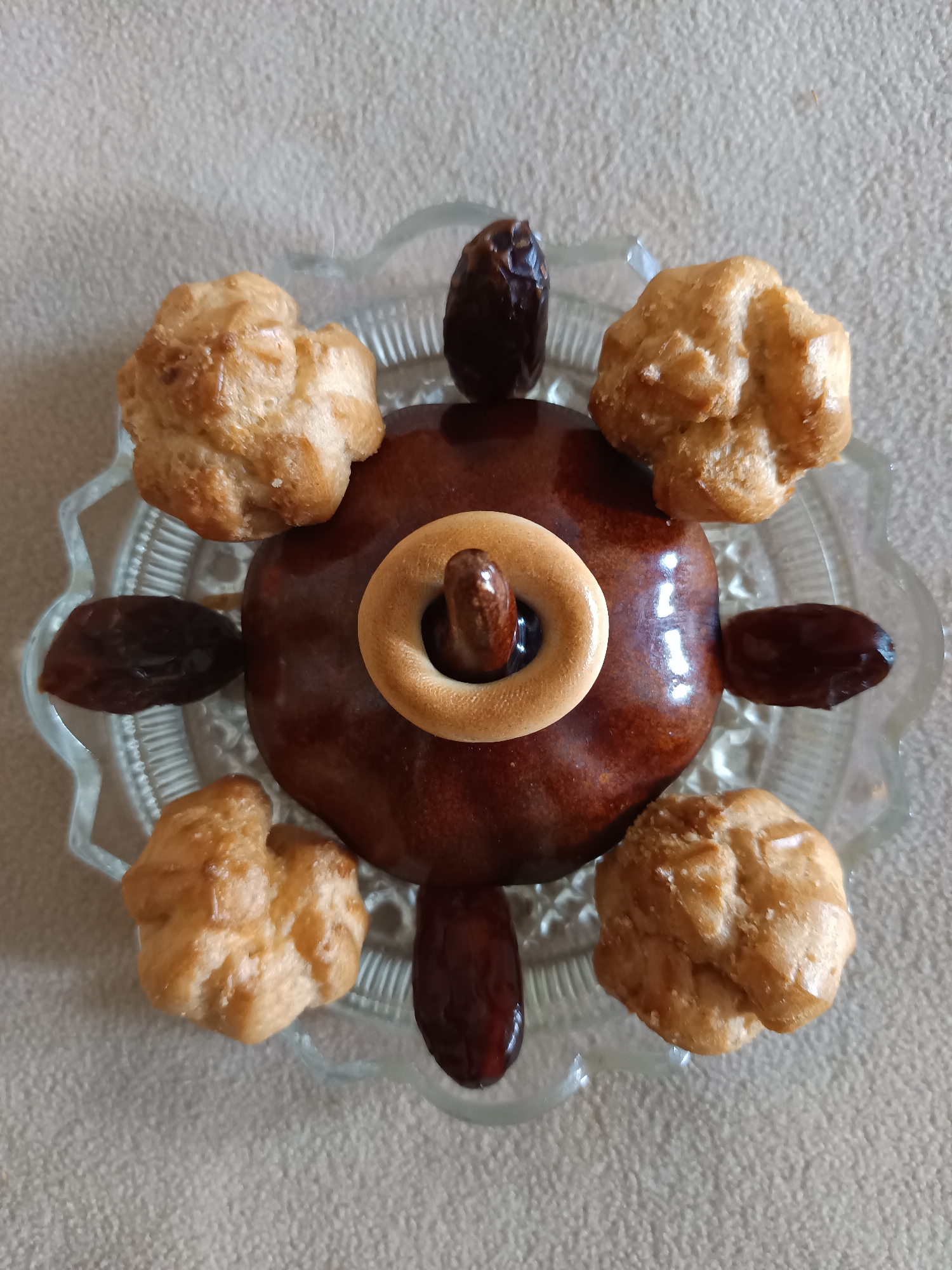 This media file is a top-down, close-up shot of a dessert arrangement on a clear, scalloped glass plate. The plate sits on a textured, neutral-colored surface, possibly a tablecloth or countertop. The main subject is a large, round, dark brown cake or pastry, possibly a cake pop or a small frosted cake, with a high-gloss finish. Placed in the center of this main pastry is a small, light brown ring-shaped cookie, possibly a mini bagel or a biscotti, with a dark brown date resting vertically within its opening.
Arranged radially around the central pastry are five small, golden-brown, puffed pastries, resembling profiteroles or cream puffs, with irregular, craggy surfaces. Between these puffed pastries, also arranged radially, are five dark brown dates.
The overall arrangement resembles a stylized flower or a celestial body with radiating petals or arms. The lighting appears to be natural and soft, casting subtle reflections on the glossy cake. There is no text visible in the image. The scene depicts a prepared dessert, suggesting it is either a snack, a small treat, or part of a larger dessert presentation. The activity is passive, focused on the static arrangement of the food items. No people are visible. The context is a domestic or food-related setting.
