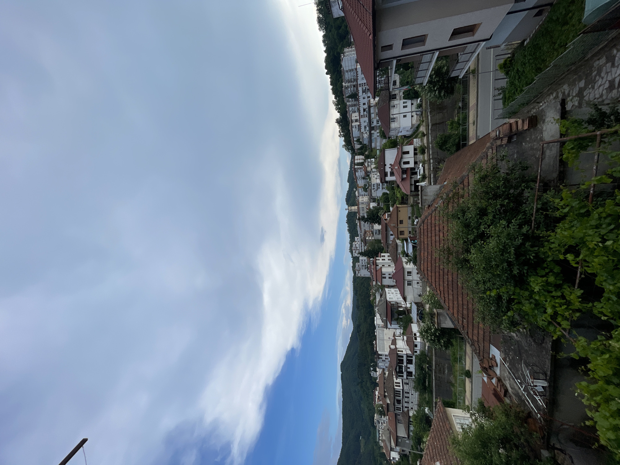 This is a high-angle, full shot of a European town, likely Zlatograd, Bulgaria, on a cloudy day. In the foreground, a red-tiled roof is visible, with some broken tiles and exposed underlayment. To the right, a portion of a modern apartment building with balconies is present. The midground reveals a densely packed residential area with buildings of varying architectural styles, predominantly with red-tiled roofs. Many of these structures feature traditional balconies and white or light-colored facades. A road with a pedestrian walkway runs horizontally through the midground. The background is dominated by lush green, tree-covered hills. A tall, white, church-like structure with a spire is visible among the buildings on the left side of the midground. The sky is partly cloudy, with patches of blue sky visible through the white clouds. No people are visible in the image.