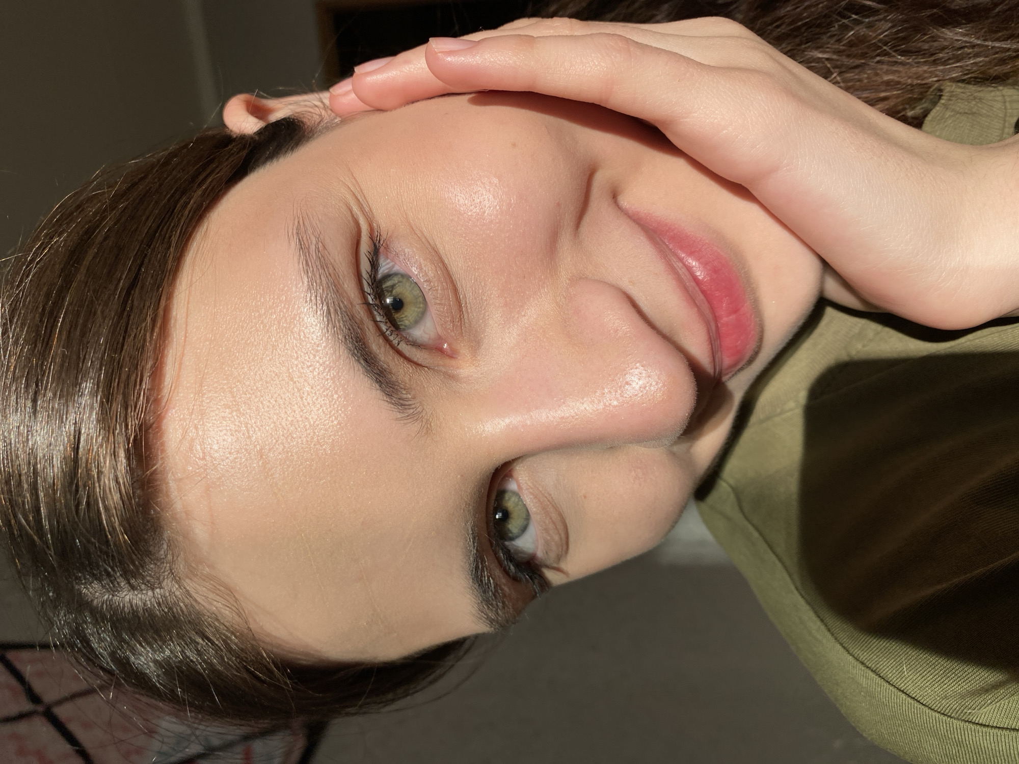 A close-up portrait of a woman with light skin and hazel eyes, her face tilted towards the right. She has dark brown hair and wears a green shirt. Her right hand is placed on her forehead. The lighting is bright and casts shadows on her face and clothing. The location is not identifiable as Votkinsk, Russia, based on the visual information provided.
