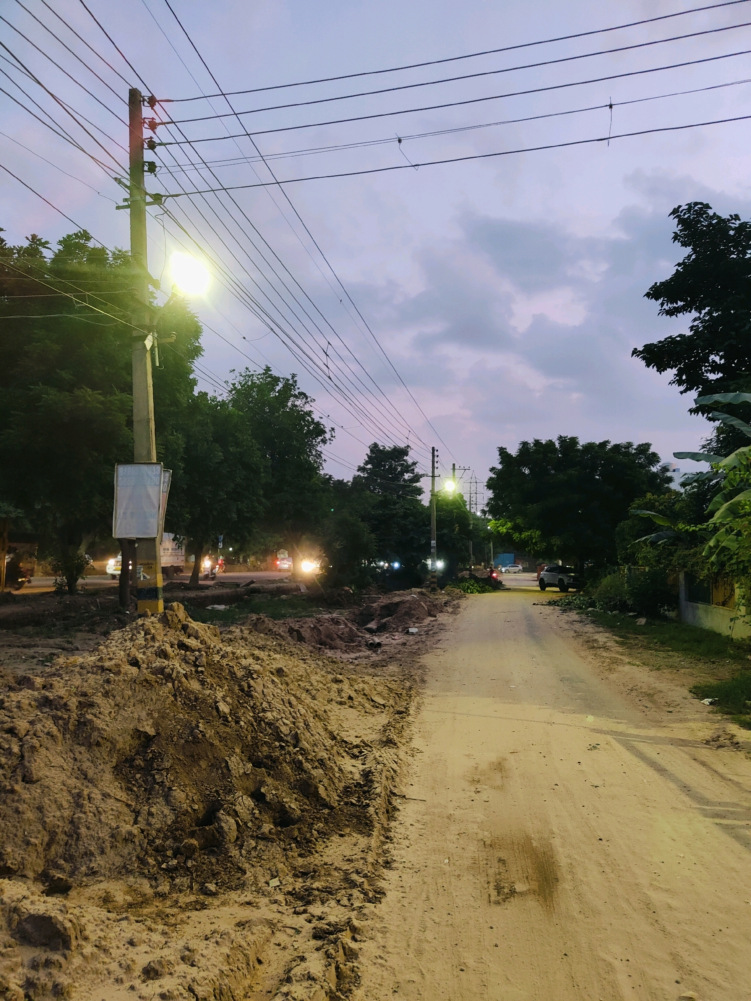 This is a photograph taken at dusk on a rural road in Gurgaon, India. The road is unpaved and dusty, with a pile of excavated earth on the left side. Overhead, power lines stretch across the sky, supported by a wooden utility pole with a street lamp that is illuminated. The sky is a muted purple and grey, with scattered clouds.
Trees line the sides of the road, and in the background, there are signs of human activity. A truck and several cars are visible on the road, their headlights on, suggesting traffic. The overall impression is one of a developing area, with infrastructure in place but the surrounding landscape still largely natural.