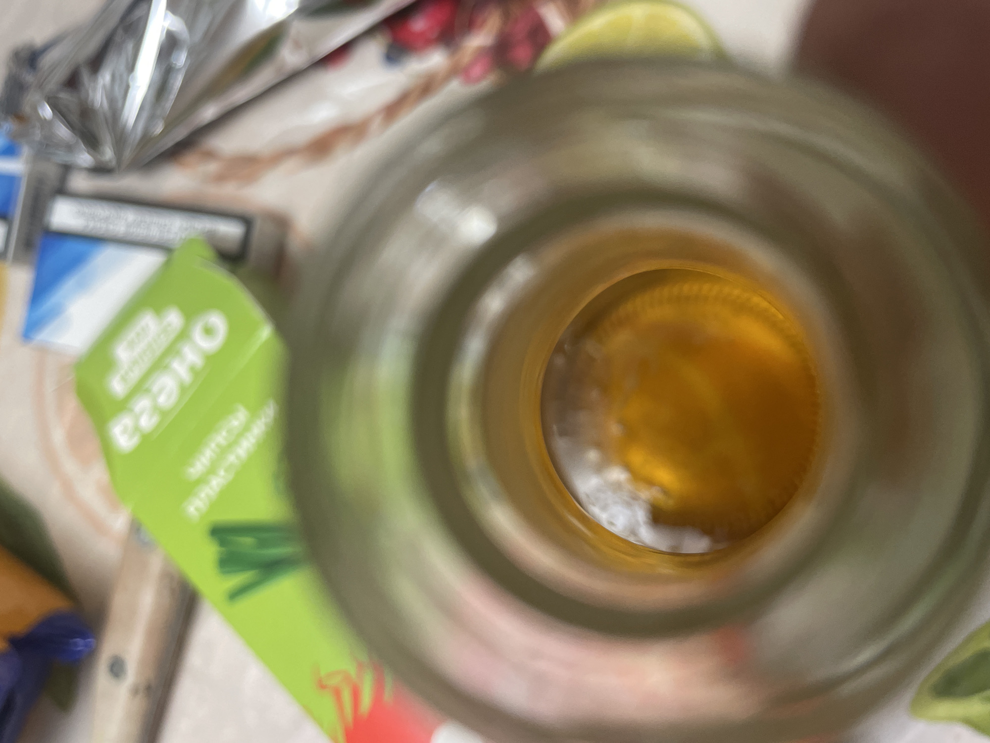 A close-up, top-down shot focuses on the opening of a glass bottle containing a yellowish liquid. The bottle's opening is circular and the interior is out of focus, with the liquid appearing slightly frothy at the surface. The background is blurry and consists of various objects, including a green package with white text, what appears to be a cigarette pack, and a crumpled silver wrapper. A wooden object, possibly a knife handle, is also visible in the upper left corner. The overall scene is indoors, likely on a flat surface like a table, and the lighting is dim. There is no indication of the location being Laishevo, Russia.