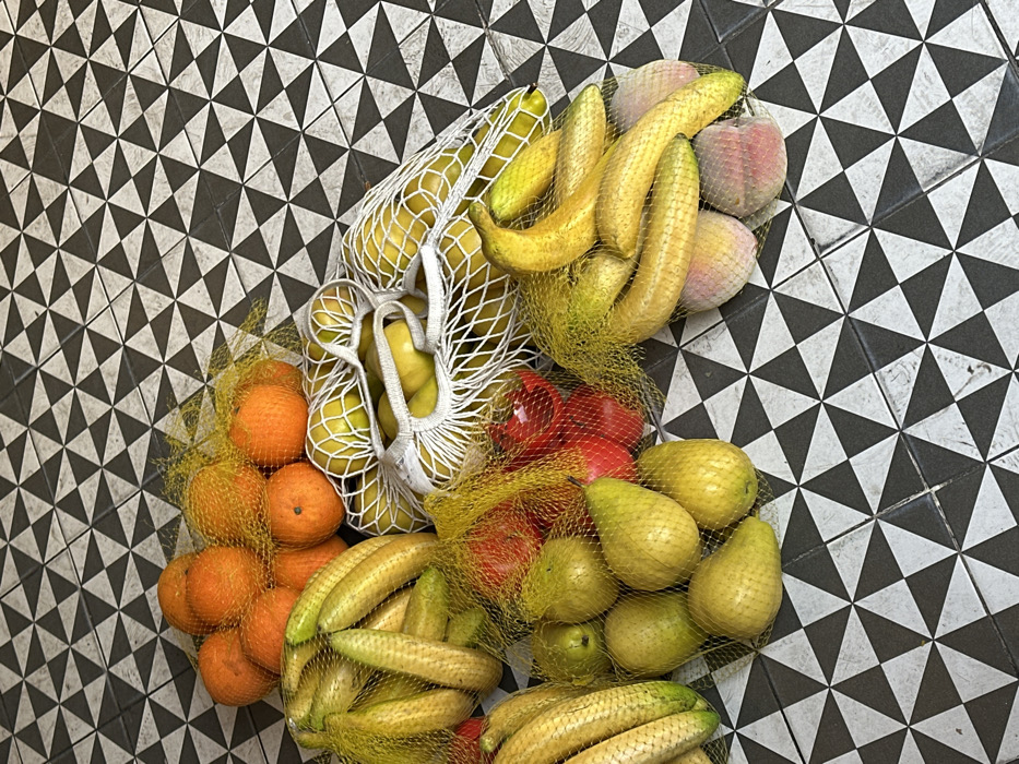 The specific location within Tel-Aviv, Israel, is not discernible from the image, though the distinctive black and white tessellating triangular floor tiles are characteristic of certain residential or commercial ground-level spaces in the city.
The scene presents an overhead view of various fresh produce items, predominantly contained within mesh bags, laid out on this patterned tiled floor. Identified subjects and objects include:
* A cluster of approximately 10-12 oranges, contained within a yellow mesh bag.
* Multiple bunches of yellow bananas, totaling an estimated 15-20 individual bananas, distributed across three separate yellow mesh bags. One of these banana bags also contains two light pink-skinned stone fruits, likely peaches or nectarines.
* Approximately 6-7 light green-yellow pears, held within a yellow mesh bag.
* A small grouping of 3-4 red bell peppers, secured in a red mesh bag.
* Three white string-net bags, each containing a mix of approximately 4-6 green apples or spherical yellow-green fruits, along with several elongated yellow fruits resembling lemons or small melons.
All produce appears fresh and intact. The items are stationary on the floor. No individuals or additional environmental context are visible.