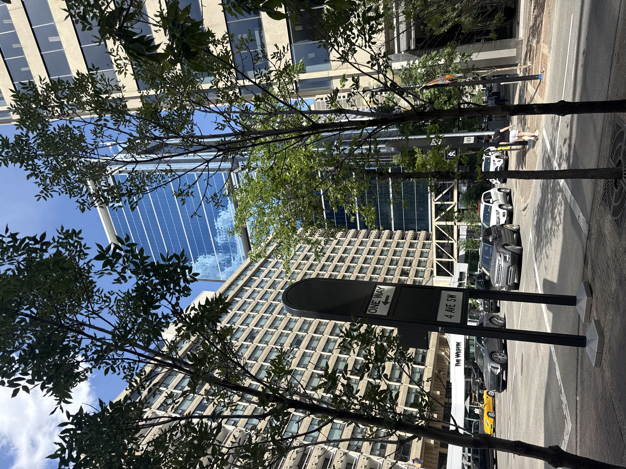 The image shows a street scene in Calgary, Canada, likely in the downtown core, with modern buildings and vehicles. In the foreground, trees with green leaves partially obscure the view of a street and sidewalk. A street sign is visible with "ONE WAY" and an arrow pointing left, as well as "4 AVE SW". Several vehicles are on the street, including a yellow sports car, a grey Cadillac SUV, a grey Range Rover SUV, and other cars. Buildings line the street, one with a beige, repetitive facade of windows, and a taller, modern building with a glass facade reflecting the sky and clouds. A person is walking away from the camera on the sidewalk. The sky is blue with some white clouds, indicating a sunny day.