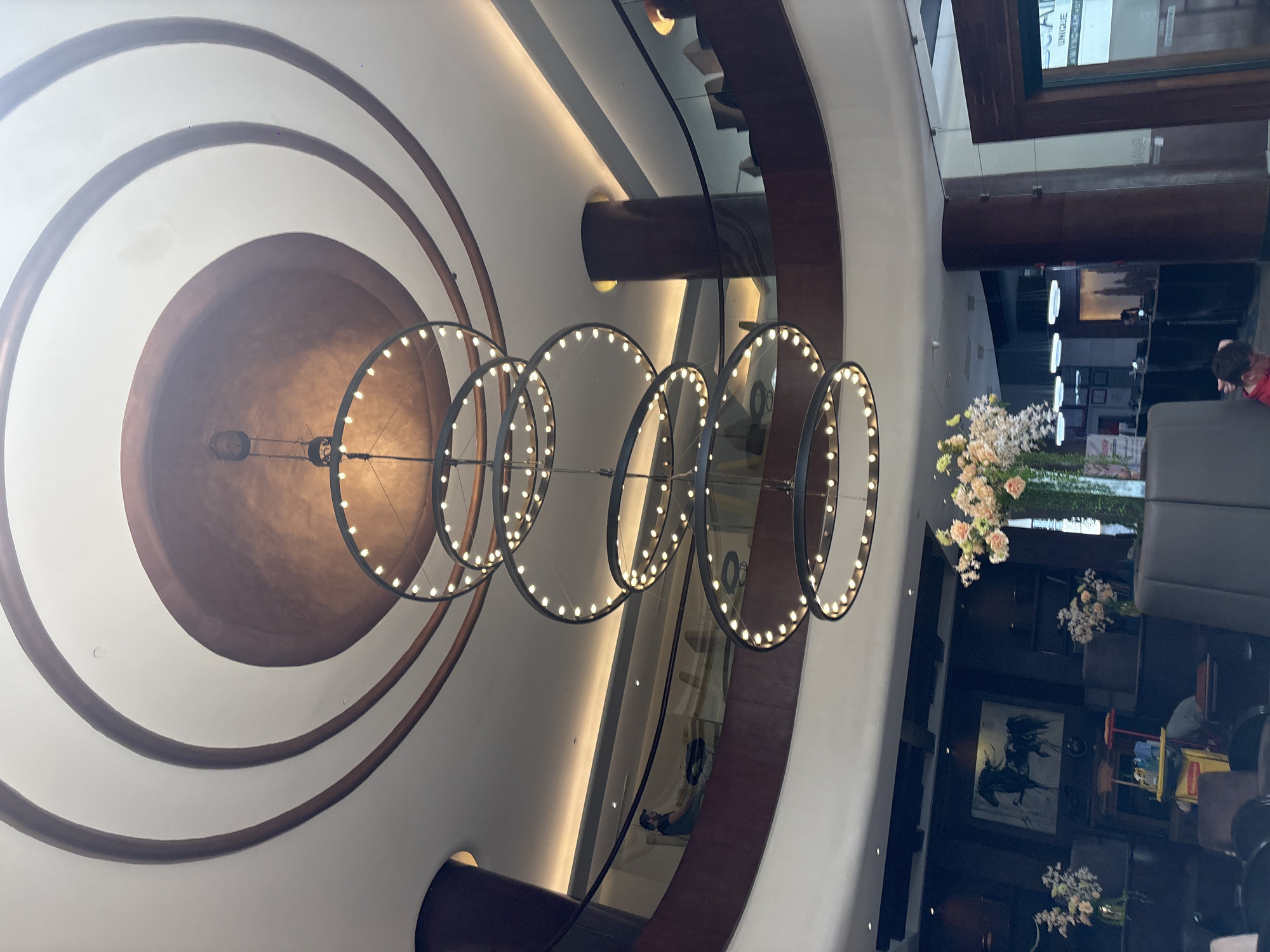 The image depicts the interior of the Gran Hotel Ciudad de México in Mexico City, Mexico. Visible is the hotel's iconic Art Nouveau ceiling featuring concentric circles of brown and white, with a modern chandelier of interconnected illuminated rings suspended from the center. Down below, floral arrangements are seen atop dark tables, with hints of seating areas and a glimpse of the hotel's lobby or common space. People can be seen in the distant background.
