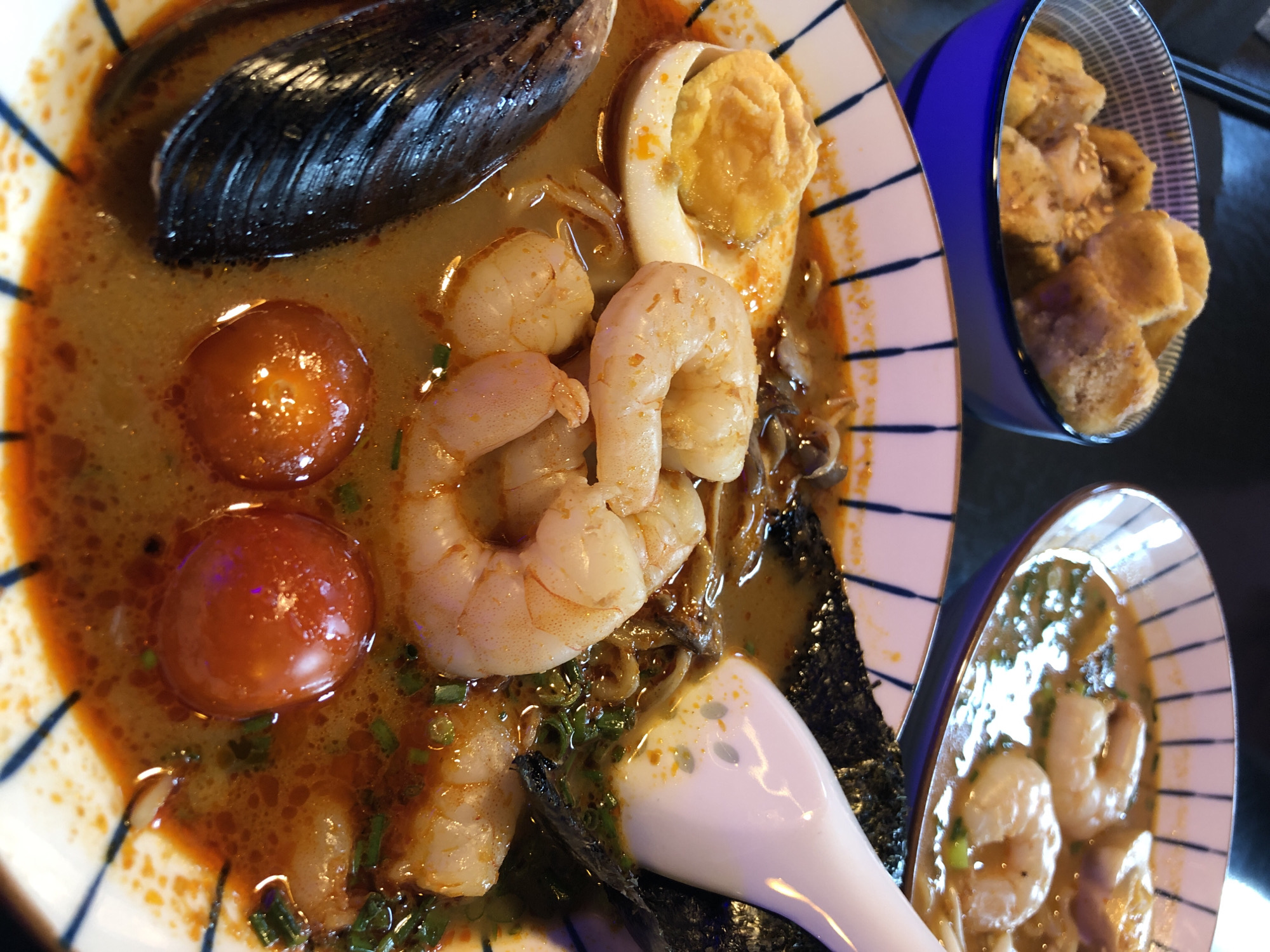 This is a close-up, eye-level, indoor shot of two bowls of ramen. In the foreground, a bowl of ramen contains a rich broth with several large shrimp, half a hard-boiled egg, a mussel shell, two cherry tomatoes, seaweed, and chopped green onions. A white spoon is resting in the bowl, partially submerged. In the background, another bowl of ramen is visible, containing shrimp and broth. To the left, a small blue and white patterned bowl holds fried morsels, possibly croutons or fried dough. The lighting is warm and appears to come from above. The scene is likely in a restaurant setting. The image does not provide any information about the city of Izhevsk, Russia.