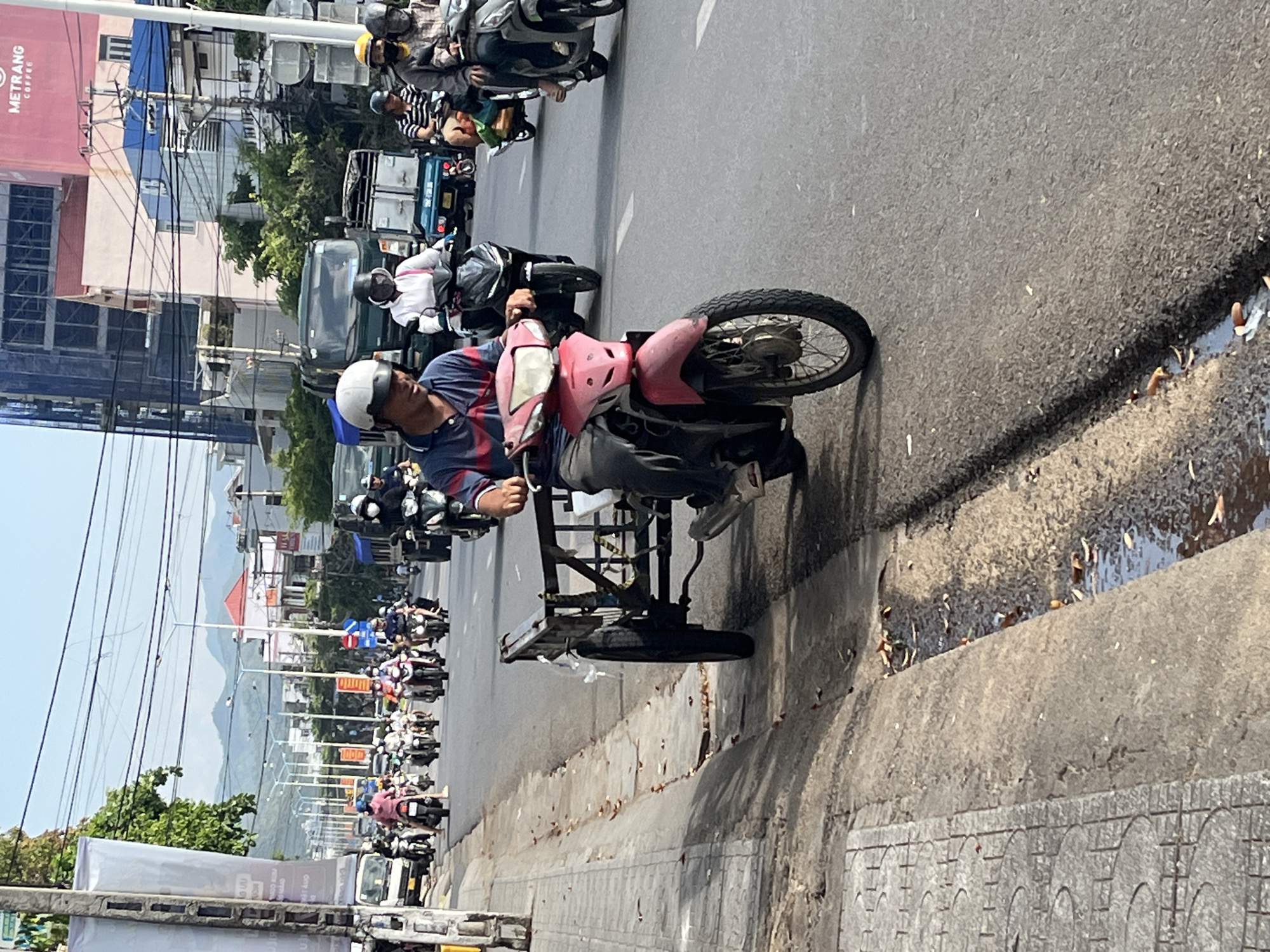 A man with a white helmet rides a three-wheeled motorized vehicle on a city street in Bac Nha Trang Ward, Vietnam. He is wearing a blue striped shirt, gray pants, and gray shoes. The vehicle is red and black with a flat bed behind the driver. The street is full of other motorized vehicles and pedestrians. The Metrang Coffee building is visible in the background.