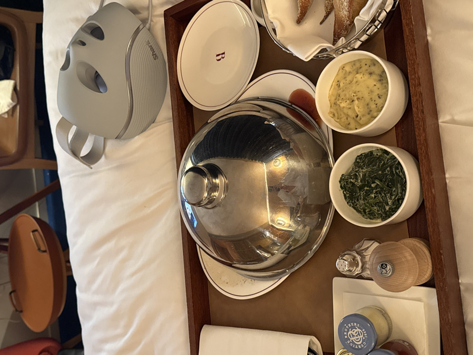 A room service meal on a wooden tray rests on white bedding in a room located in Unknown, Unknown. The tray features a large white plate covered by a silver domed cloche, and an oval white plate with a red "B" logo. Two white bowls contain creamed spinach and mashed potatoes. A small metal basket holds several fried, sugar-dusted pastries. A white condiment plate displays two glass jars, a salt shaker, and a wooden pepper mill. A folded white napkin is also present. Adjacent to the tray on the bedding is a light grey "Shark" branded facial mask device with an attached strap and cable. In the background, out of focus, are wooden furniture elements and an orange, circular side table with handles.