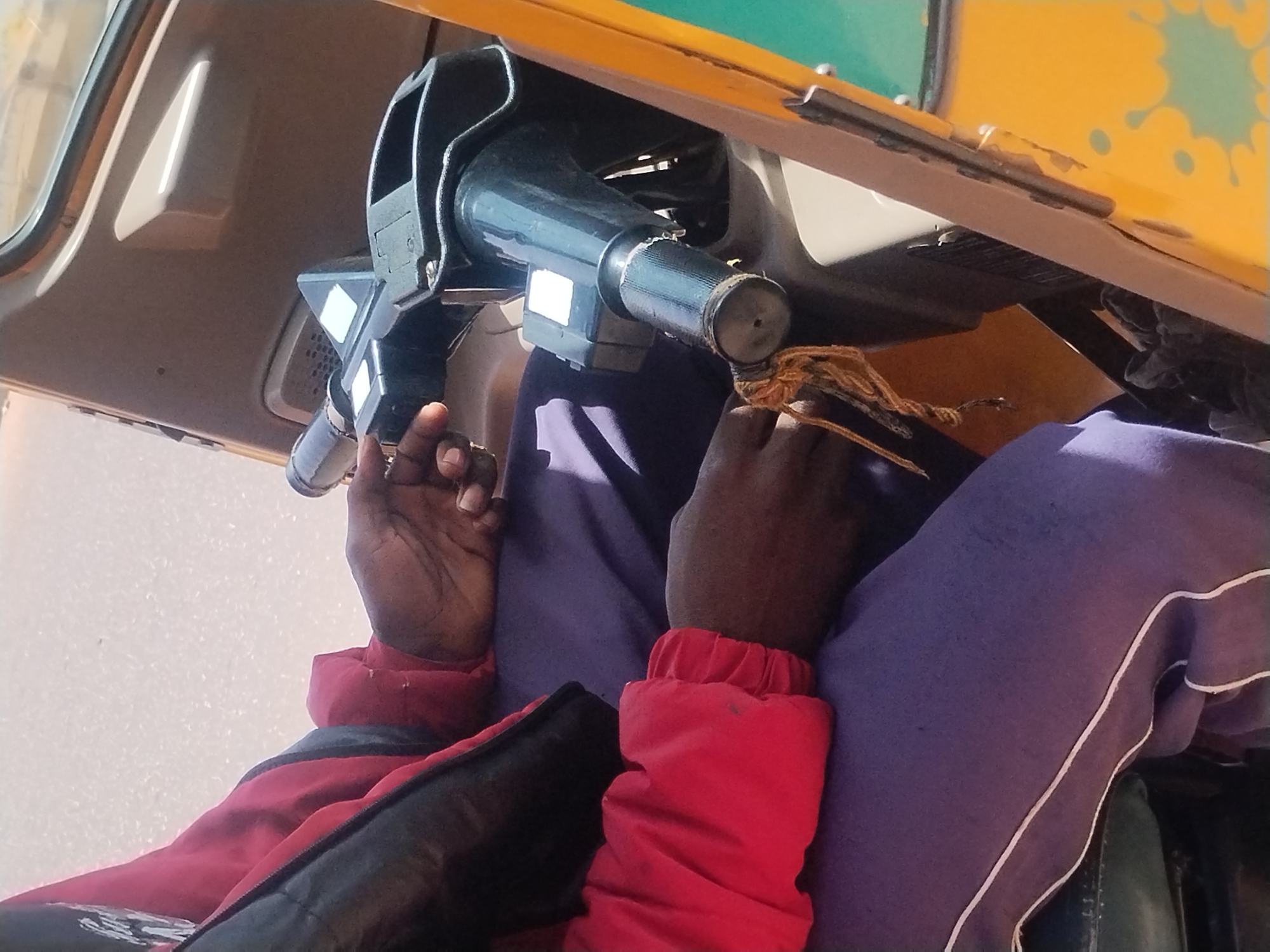 A low-angle, medium shot, taken in the city of Jos, Nigeria, shows a person holding the steering mechanism of an auto-rickshaw. The auto-rickshaw is painted yellow with green accents. The person has dark brown skin and is wearing a red jacket with black accents over purple pants with a white stripe. Their hands are gripping the black, cylindrical steering column. A piece of knotted brown string is tied to the steering mechanism near their right hand. The background is a mix of light and dark colors.