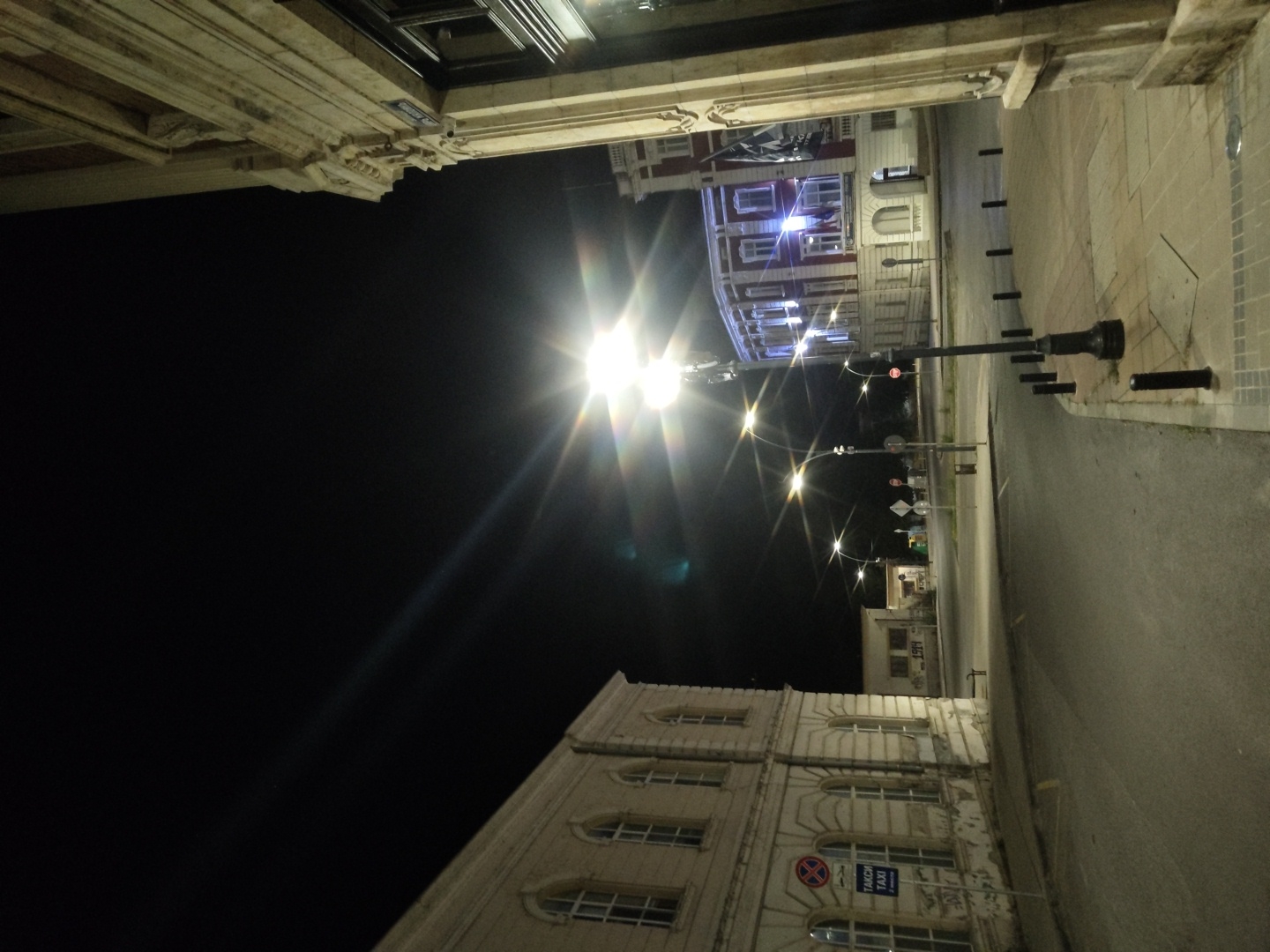 The image shows an intersection at night in Ruse, Bulgaria. The street is paved and empty, with streetlights illuminating the scene. On the left is a large, pale building with arched windows. A blue sign with "TAKSI TAXI" is visible on its facade. Across the street is a red and white building with illuminated windows. The sky is dark, and the streetlights create bright starbursts of light. There are no people or vehicles visible in the image. The overall impression is a quiet, deserted urban landscape at night.