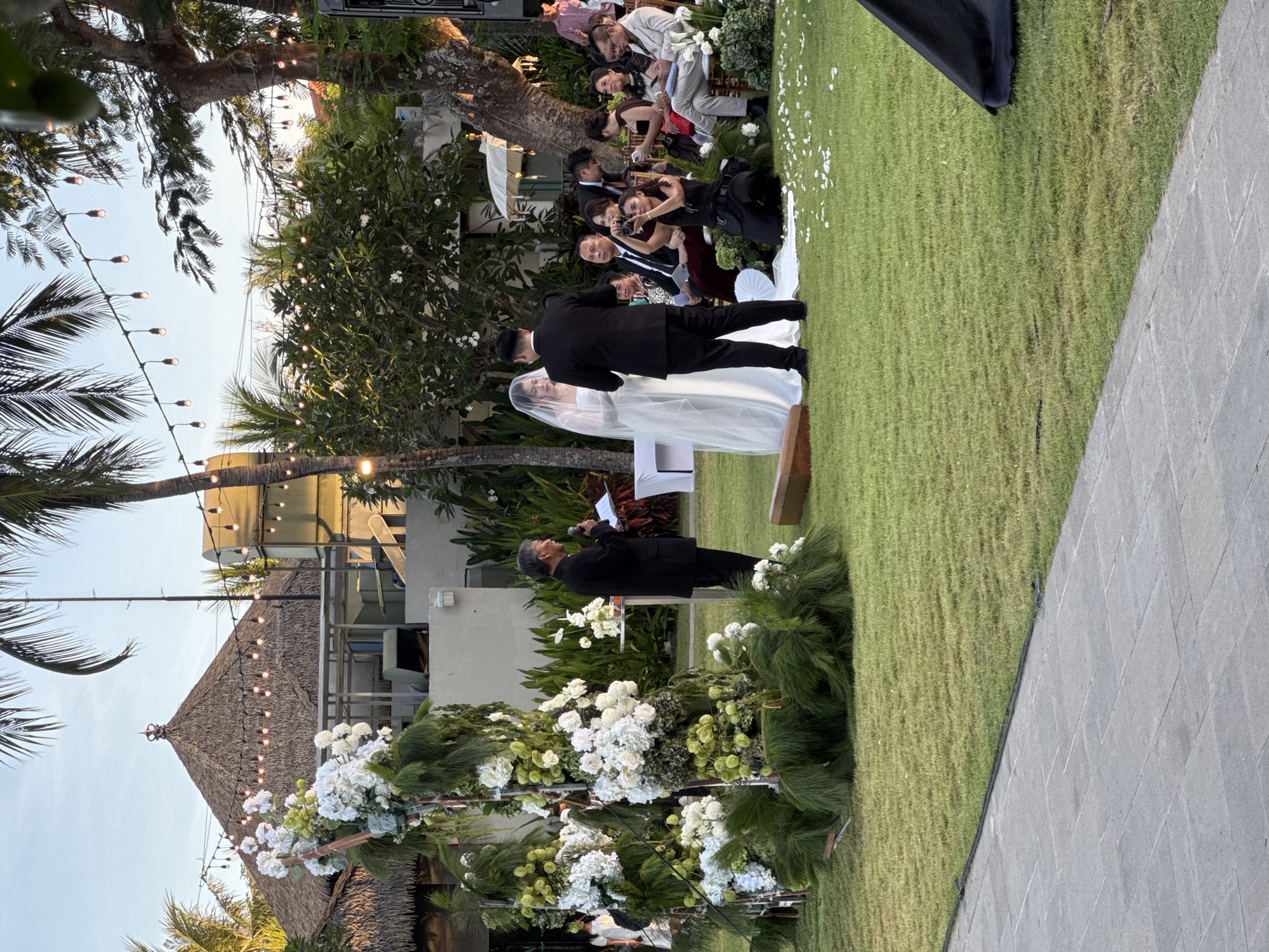 Here is a description of the image:
The image shows a wedding ceremony taking place outdoors on a grassy lawn, presumably in the city of Unknown, Unknown. A bride and groom are standing together, seemingly during their vows or exchanging a kiss, with the officiant standing to their side, reading from a paper. Guests are seated in the background, watching the ceremony. Some guests are taking photos. The setting includes tropical plants, string lights, and a building with a thatched roof. A decorated archway with white flowers adorns the foreground.
