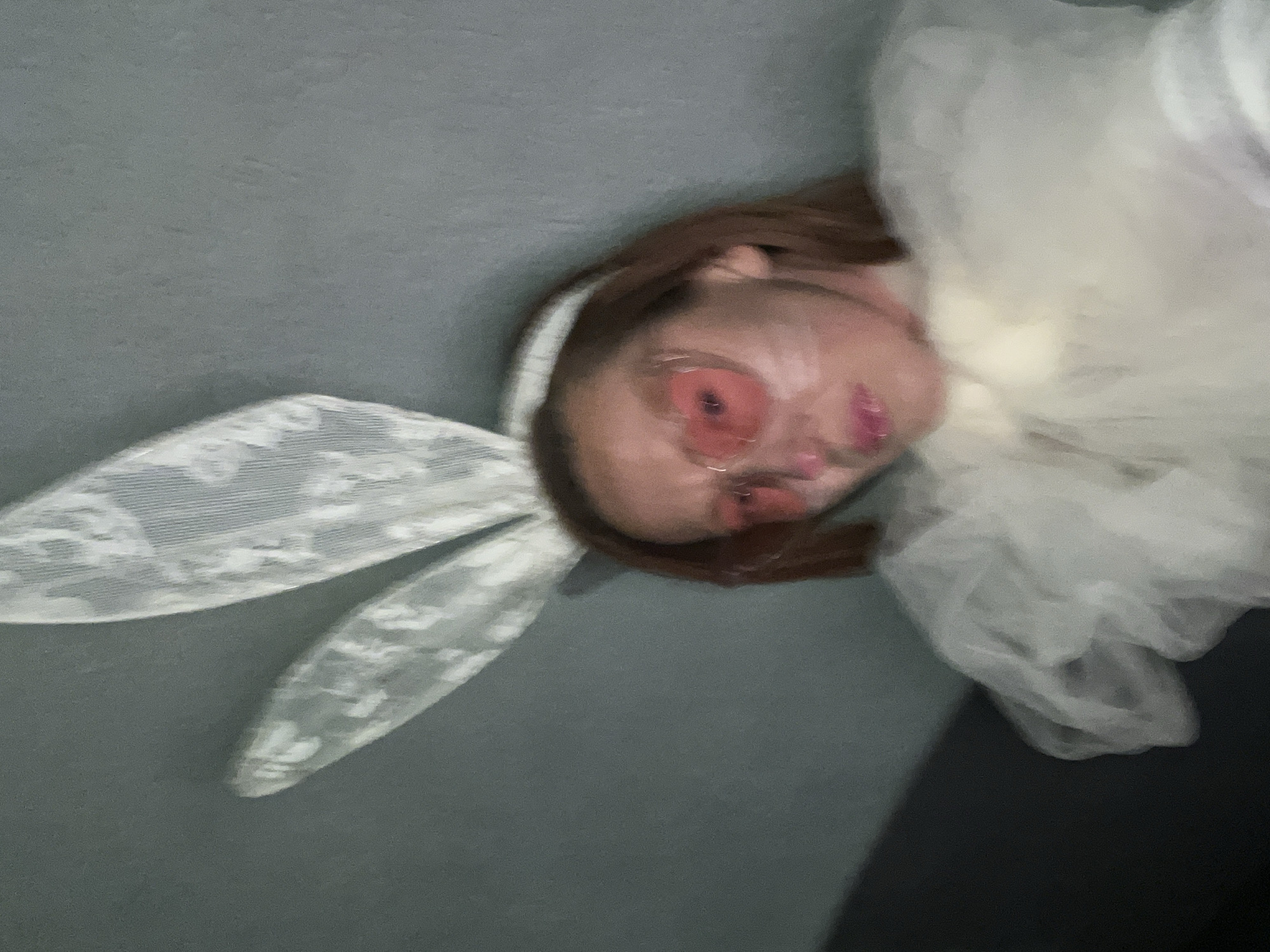 A blurry, tilted close-up image shows a person wearing lace bunny ears and what appears to be a white tulle dress or fabric. The person's face is visible, featuring red-tinted glasses and pink lipstick. The background is a solid gray color in the top left and lower right areas, suggesting a floor or wall. No identifiable landmarks or information is present to indicate that the scene is located in Budva, Montenegro. The environment appears to be indoors.